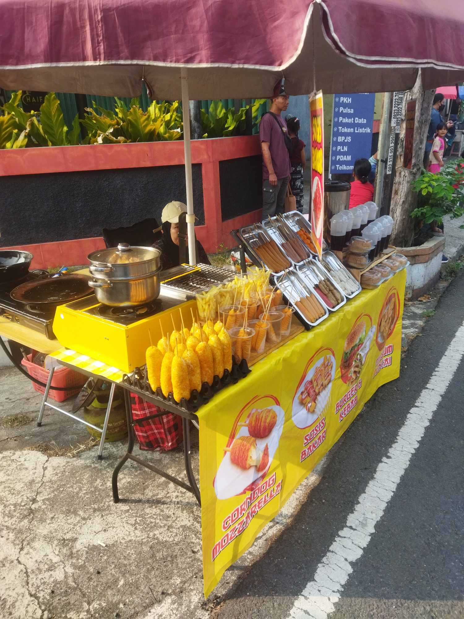 A street food vendor stall with a yellow tablecloth is set up on a paved area next to a road in Wonogiri, Indonesia. The stall displays a variety of food items, including corn dogs, sausages, and what appear to be French fries. A woman wearing a headscarf is partially visible behind the stall, possibly attending to customers. In the background, several people are standing, and a man is looking at a sign with text. The scene is illuminated by sunlight, casting shadows on the ground.
