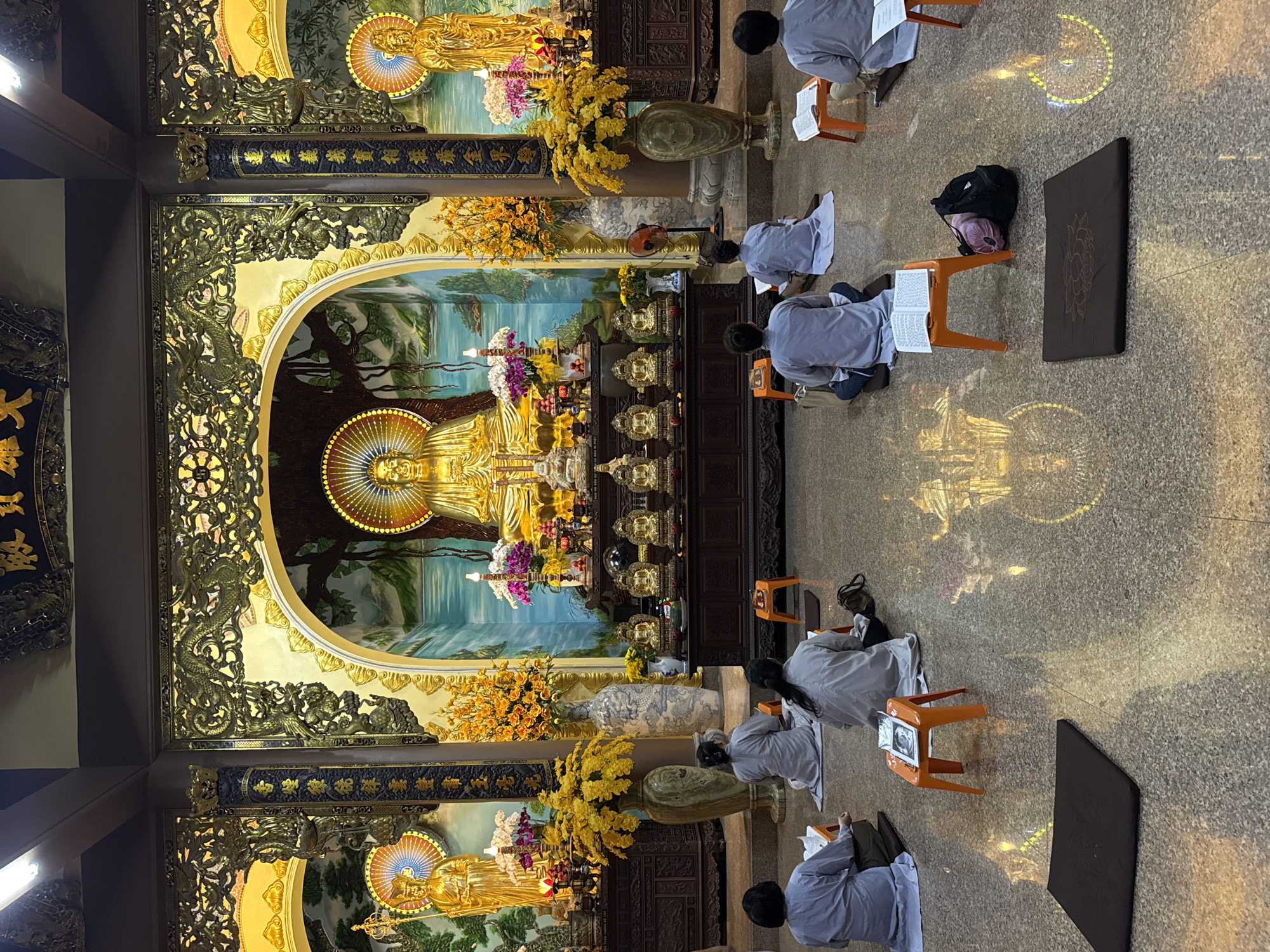 The image depicts a Buddhist temple in Đà Nẵng, Vietnam. Within the temple, several individuals dressed in light gray robes are engaged in prayer. Each person is seated on the floor, with an orange stool in front of them holding a book or scripture. A black backpack sits near one of the individuals. The temple's architecture is ornate, featuring gilded statues of Buddha set against backdrop scenery. The floor is tiled and reflects the light and colors of the temple. Yellow floral arrangements adorn the space around the statues.