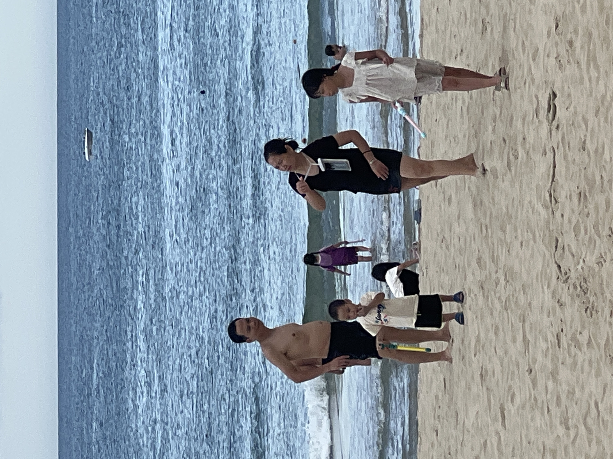 The image depicts a group of people on a beach in Da Nang, Vietnam. The scene is set with the ocean on the left and a sandy beach on the right. The water is a deep blue, indicating it could be daytime, and the sky appears overcast. Several individuals are visible, including adults and children, engaged in various beach activities.
In the foreground, a shirtless man in black shorts stands facing the water. To his right, a young boy in a white t-shirt and black shorts is also present, holding a yellow object that resembles a toy water gun. Further back, a woman in a black dress and shorts is seen from the side, gesturing with her hand and looking down. Beside her, a young girl in a white dress with a ruffled collar is looking towards the ground. In the mid-ground, another person in a purple outfit is visible. In the background, more figures can be seen, and what appears to be a black and white umbrella or shelter on the sand.
The overall mood seems relaxed and casual, typical of a beach outing. The presence of toy water guns suggests children are actively playing. The composition of the image, with the ocean and sand clearly delineated, along with the various figures scattered across the beach, creates a sense of a lively and communal environment. The lighting is diffused, consistent with an overcast day. No specific time of day can be definitively determined, but it is likely daytime. No visible text is present in the image.