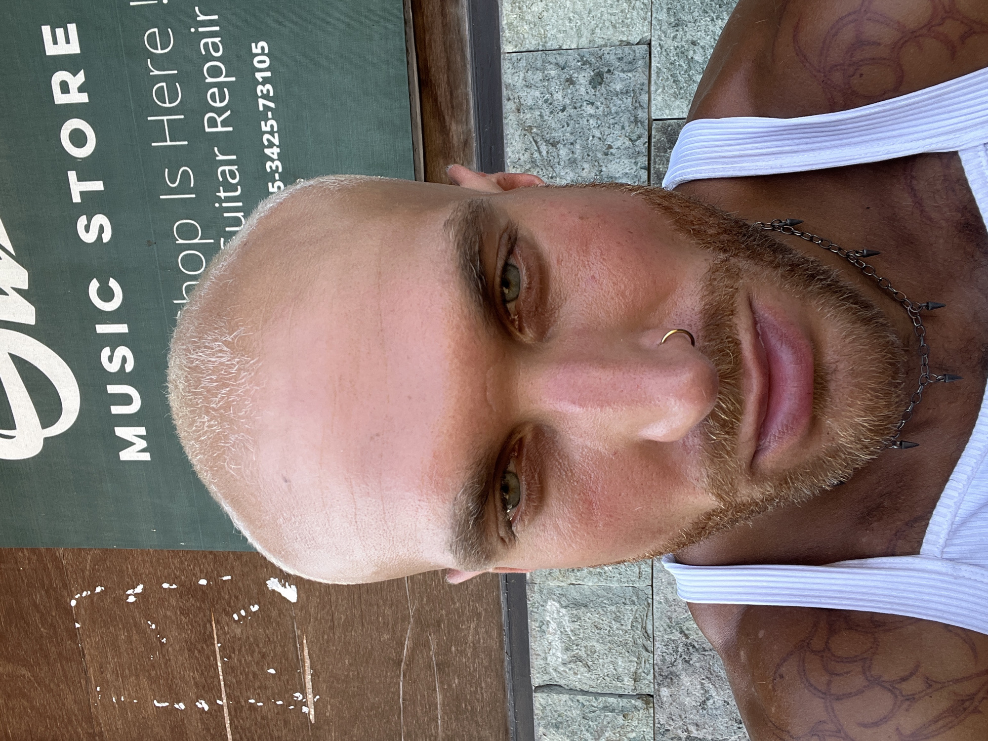 The image is a close-up, front-facing selfie of a bald, fair-skinned man with light brown hair and beard. He is wearing a white ribbed tank top, a gold hoop nose ring, and a chain necklace with spiked pendants. He has tattoos on his left shoulder and upper arm. The background consists of a stone wall, a wooden surface, and a green sign with white lettering that reads "MUSIC STORE" and "Shop Is Here! Guitar Repair 5-3425-73105". The lighting suggests it is daytime, possibly outdoors. The location is specified as North Kuta, Indonesia, although this context is not visually evident within the image itself.