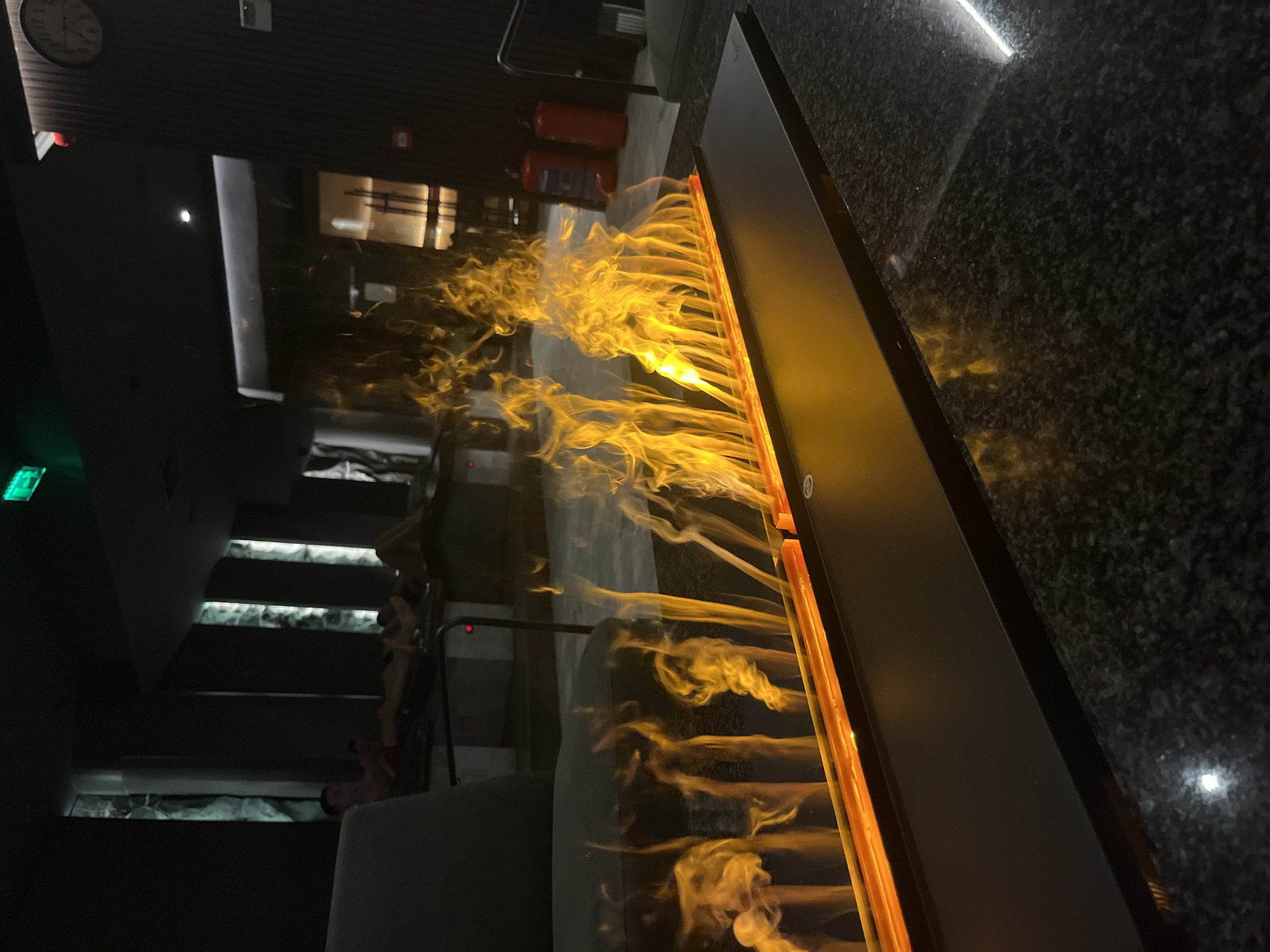 The image shows an indoor scene, possibly a commercial establishment, in Sofia, Bulgaria. A long, rectangular, modern electric fireplace with simulated orange flames dominates the right side of the frame. The fireplace sits on a dark granite surface. In the background, there's a set of stairs with illuminated treads, possibly leading to a lower level. People are visible on the stairs. Further back, there's a corridor or hallway. A clock is mounted on the wall at the top left. Red fire extinguishers are visible in the background. An exit sign is visible at the top left.