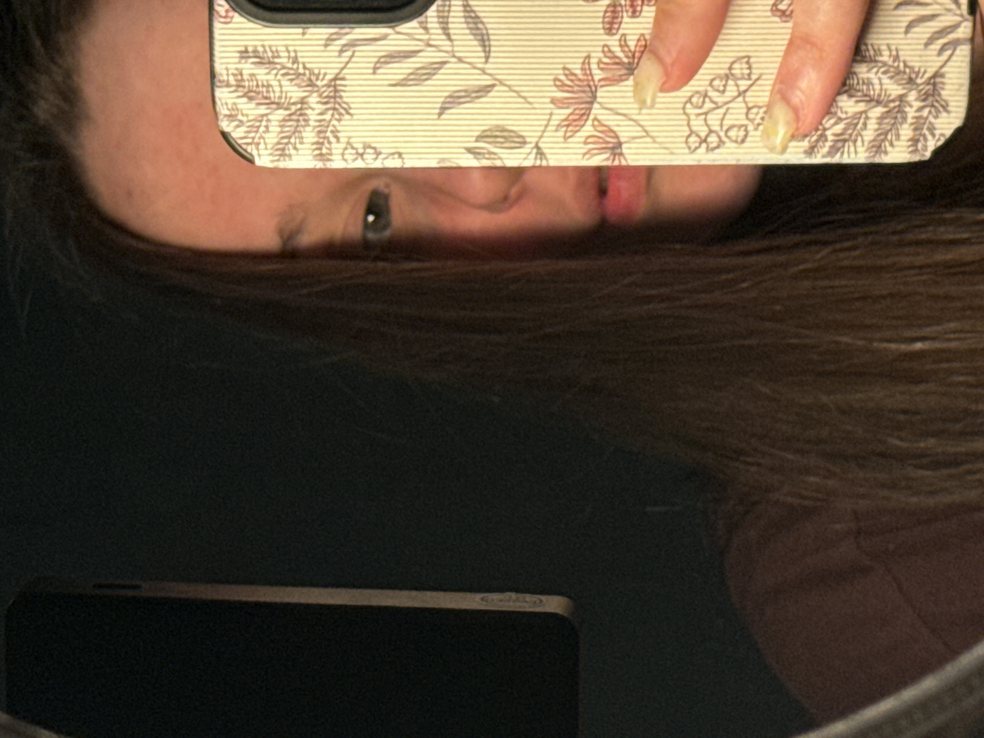 A woman with long brown hair is taking a mirror selfie. Her face is partially obscured by the phone in her hand, which has a floral patterned case. The woman has an eye visible and her lips are pursed slightly. A second phone is visible in the reflection of the mirror. The background appears to be a dark, textured surface. There is no information to determine the location to be Krasnodar, Russia.
