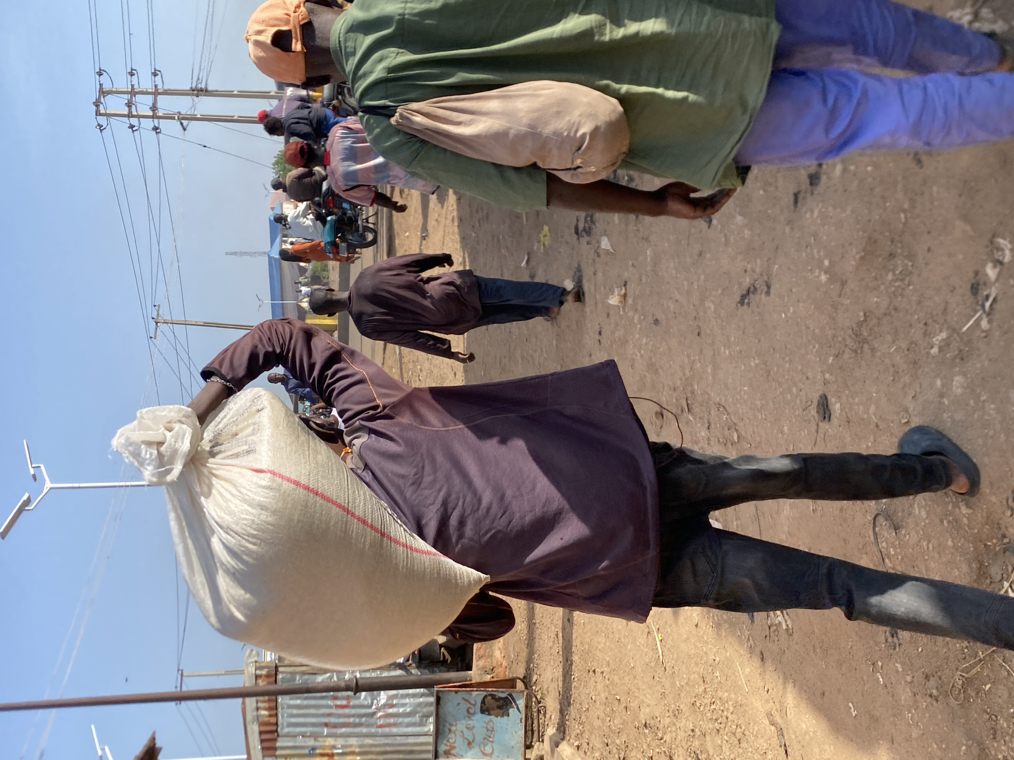 In Bukuru, Nigeria, a man is captured from behind, carrying a large sack of grain over his shoulder. He is dressed in a dark purple long-sleeved shirt and dark denim jeans, with dark slip-on shoes. The sack is made of a light-colored woven material, possibly plastic, with a red and white striped trim. The ground is unpaved and dusty, with scattered debris. In the background, other individuals are visible walking, some on foot and others on a motorcycle. Electrical poles and wires stretch across the sky, indicative of an urban or semi-urban setting. A corrugated metal structure is present on the left side of the frame, with a sign that reads "Next Level Cuts" and features a silhouette of a man with a fade hairstyle. The scene appears to be a marketplace or a busy thoroughfare.