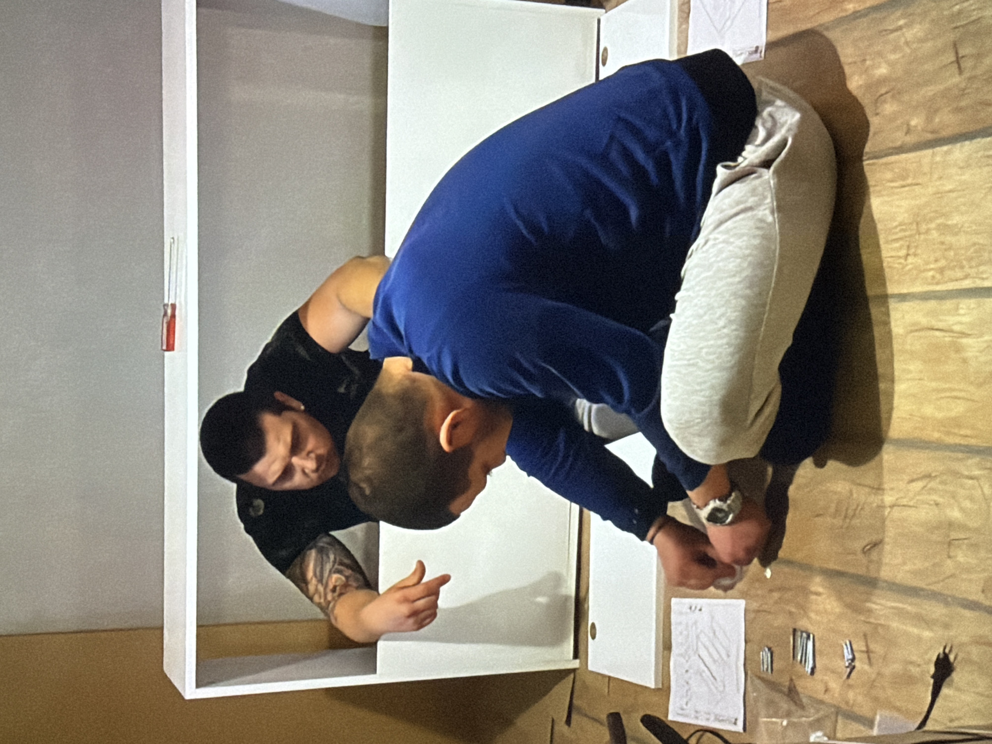 Two men are assembling furniture indoors, likely in Burgas, Bulgaria. One man, wearing a black shirt and showcasing tattoos on his arm, is positioned inside the base of a white cabinet, pointing outward. Another man, dressed in a blue sweater and grey sweatpants, is crouched over the assembly area, possibly inserting screws or other fasteners. Nearby on the floor are assembly instructions, various hardware components, and an electrical cord. The floor appears to be a wood or laminate material. A screwdriver is attached to the inside of a shelf.