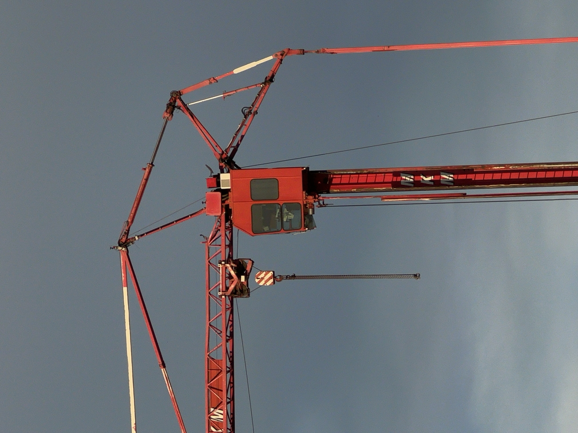 The image shows a close-up of a red tower crane against a cloudy, grayish-blue sky. The crane has a large boom extending horizontally to the left and a vertical tower structure. On the tower, a red operator's cabin with windows is visible. A chain with a hook is hanging from the boom, and a red and white striped pulley is attached to the hook. The word "MKM" is visible vertically on the tower. The scene is likely from a construction site.