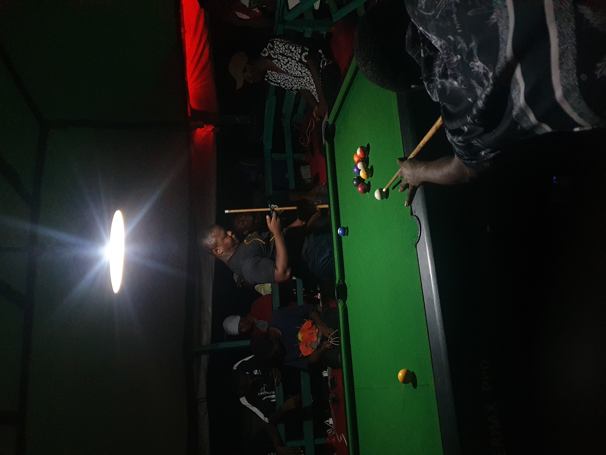 The image depicts an indoor pool hall, likely in Lokoja, Nigeria, characterized by its dimly lit ambiance, with a prominent overhead light fixture illuminating the green felt pool table. Several individuals are present, engaged in or observing a game of pool. One person, wearing a black t-shirt, stands near the center of the table, appearing to be looking at their phone. Another person, seated and facing away from the camera, is in the foreground, preparing to strike a white ball with a cue stick. Other individuals are visible in the background, some seated and others standing, observing the game. The environment appears to be somewhat informal, with red fabric visible in the upper background, suggesting a draped or tent-like structure, and wooden benches lining the area. The pool table itself is set with several colored balls arranged in a triangular formation and a single blue ball positioned on the felt. The overall scene suggests a casual gathering centered around a game of pool.