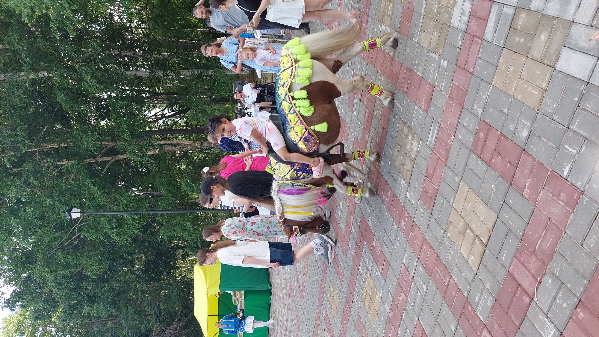 In Komsomolsk-on-Amur, Russia, a young girl is being paraded on a pony adorned with colorful tassels and bright yellow leg wraps. The pony, a mix of brown and white, has a mane and tail decorated with purple and white ribbons. The girl, wearing a light-colored outfit with a pink pattern, sits confidently on the pony's saddle, which is covered in a sequined blanket with yellow tassels.
The scene unfolds on a paved walkway made of red and gray interlocking bricks. Behind the pony, a group of people are visible, some walking and others observing. To the left, a yellow and green tent stands, suggesting an outdoor event or fair. The background is filled with lush green trees, indicating a park or outdoor recreational area. The lighting suggests it is daytime.