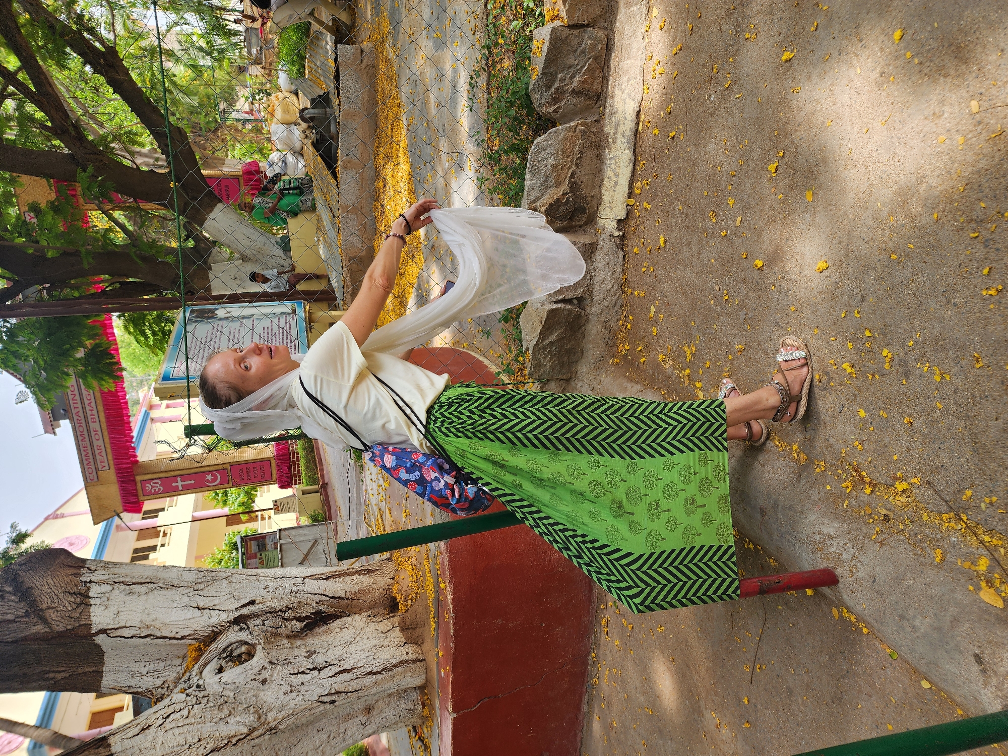 A woman stands in an outdoor setting. She wears a white headscarf, a cream-colored shirt, and a green skirt with a black chevron pattern at the edges and tree-like designs in the center. She holds a translucent white scarf above her head, and a colorful purse hangs from her shoulder. She wears sandals on her feet and stands on a concrete surface covered in scattered yellow flower petals. To her left, there is a large, white-painted tree trunk, and behind her, a red and green metal structure. In the background, there are buildings with religious banners and signs in a foreign language. A chain-link fence and stone wall separate the woman from the background buildings. There is a lush green tree overhead, partially obscuring the view. The setting appears to be in a public area, possibly a park or garden adjacent to a religious site.