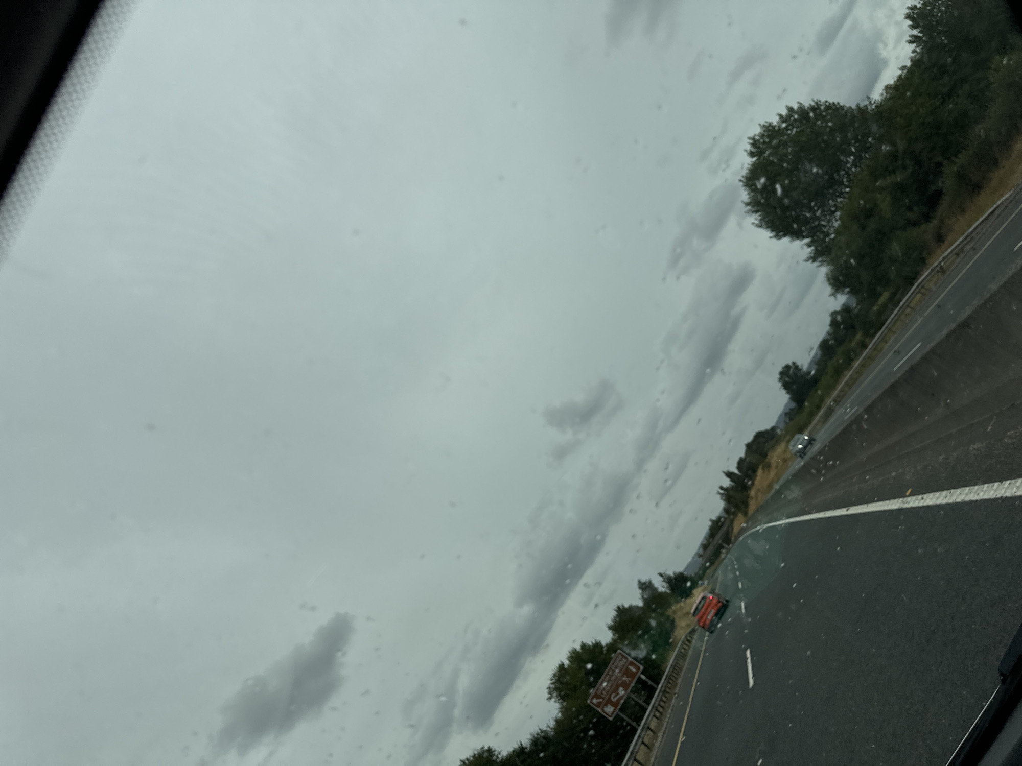 The image depicts a cloudy sky over a highway in Tinryland, Ireland. A red car is driving on the highway, with a white line marking the lane. Trees and vegetation are visible on the side of the road. There are water droplets on the windshield, suggesting it is raining. A road sign with an arrow and icons is also present.