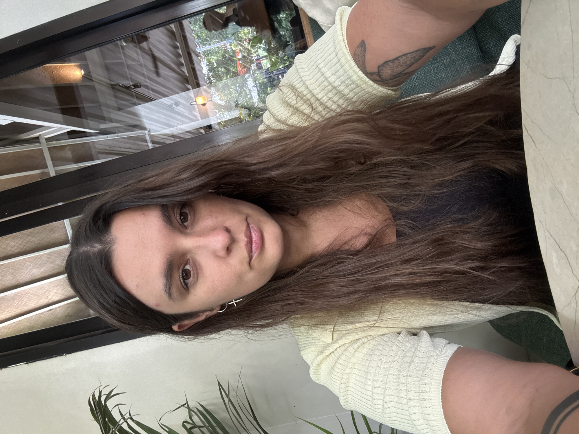 A medium shot portrait shows a woman with long brown hair in a light yellow cardigan and black top. She is seated indoors, with a window behind her that reflects an outdoor scene with greenery and building structures. A potted plant is visible to her left. The woman is looking directly at the camera.
