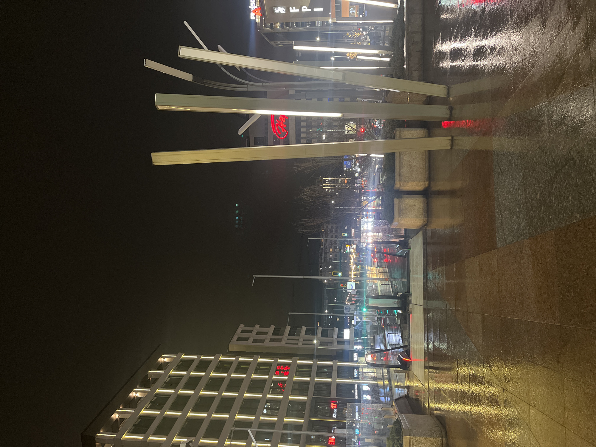 The image captures a nighttime street scene in Sofia, Bulgaria, characterized by wet, reflective pavement. In the foreground, there are several illuminated, elongated, rectangular metal structures, appearing as a modern art installation. Further in the background, there's a building facade with a grid-like pattern of illuminated windows. A red signage is visible on a building in the distance. Street lights and traffic signals dot the horizon, blurred by distance and possibly moisture in the air. Reflections on the wet ground create a distorted mirror of the light sources, including red traffic lights and white streetlights. The overall impression is of a city environment at night after a rain. The architecture and signage suggest a contemporary urban setting, although no people are clearly visible in the photo.