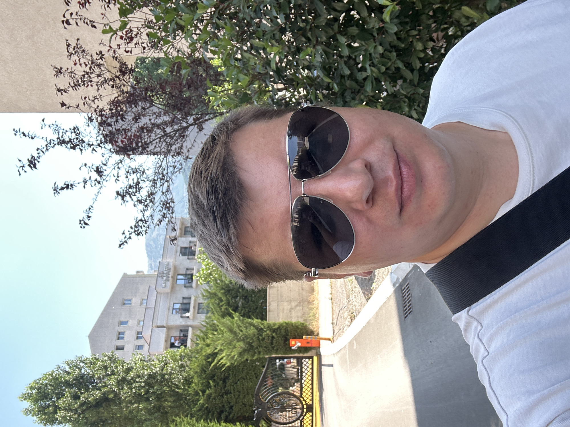 A selfie taken by a man wearing sunglasses and a white t-shirt, with a black strap across his chest. He is standing outdoors, with a building and trees in the background. The building appears to be a hotel with several balconies. The setting is in the city of Budva, Montenegro, indicated by the hotel's signage which reads "HOTEL MONTE ROJA." To the left, there is a decorative black metal gate with a bicycle parked behind it, and an orange bollard. The ground is paved with asphalt and concrete. The sky is clear and blue.