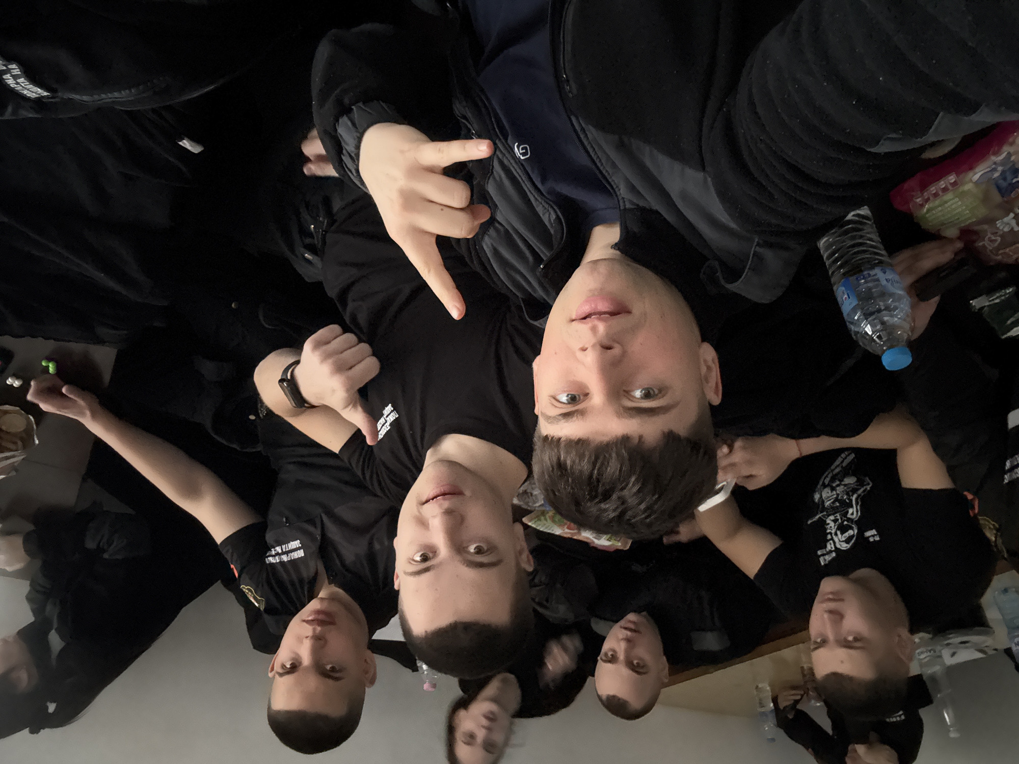 A group of six young men are posing for a selfie indoors. The location is identified as Sofia, Bulgaria. All are wearing dark clothing. One person on the upper right is holding up his hand giving the "devil horns" sign. A water bottle and snacks are visible to the right of the frame. The men are sitting around a table.