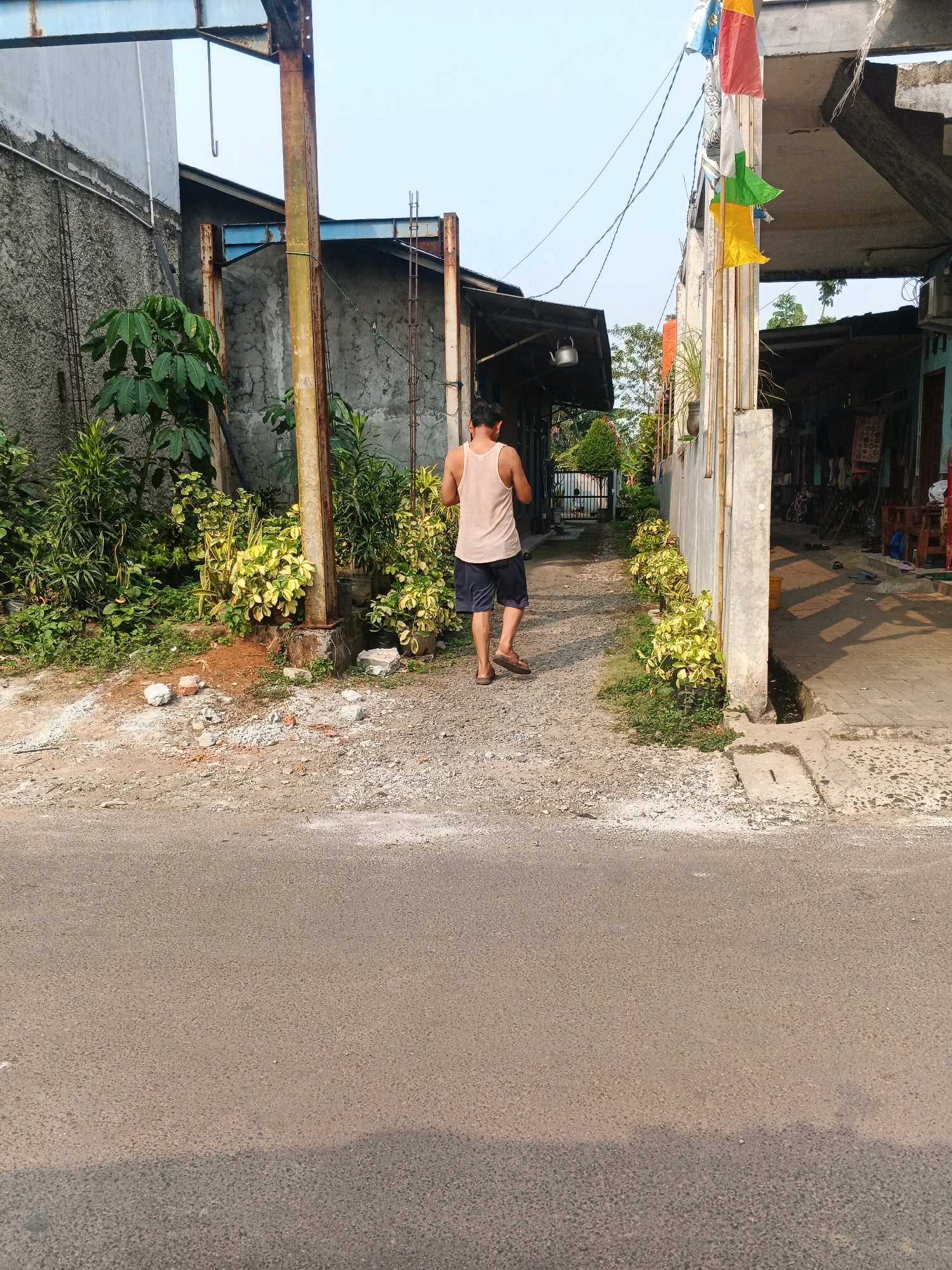 A medium shot shows a man in a light-colored tank top and dark shorts walking away from the camera on a gravel path. The path is flanked by lush green vegetation on the left and buildings on the right. Colorful flags are strung above the buildings on the right, suggesting a festive occasion. The man is wearing flip-flops. The overall scene appears to be in a residential or commercial area with a clear sky. The environment appears to be Kadu, Indonesia, based on the available information about the image.