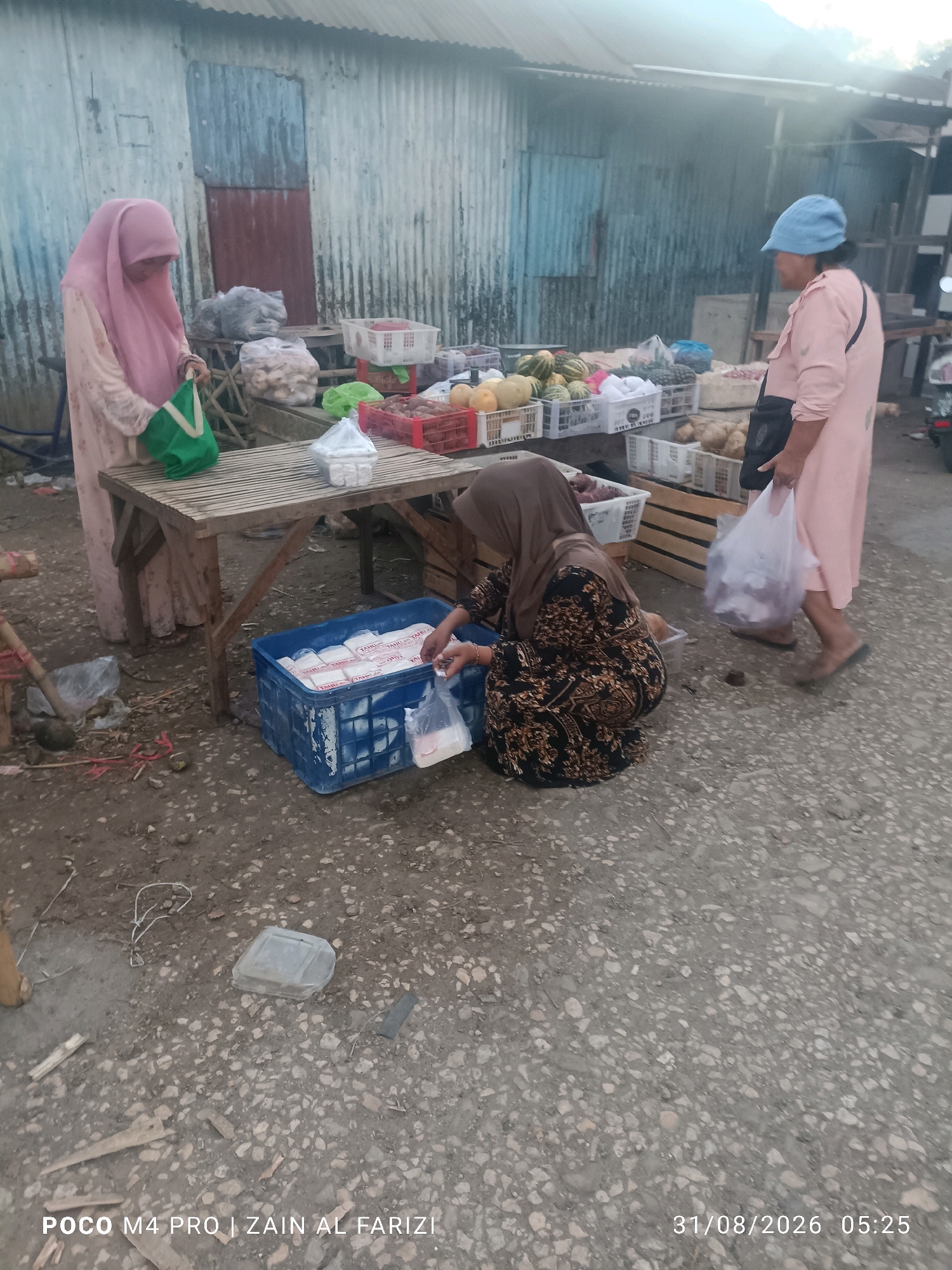 In the city of Arosbaya, Indonesia, an outdoor market scene unfolds. Three individuals are present. On the left, a woman wearing a pink hijab and a light-colored abaya stands by a wooden table, holding a green tote bag. In the center, another woman, also in a hijab, kneels beside a blue plastic crate filled with white blocks of tofu. She is in the process of bagging some of the tofu. To the right, a woman in a pink abaya and a blue cap stands looking away from the camera, holding a plastic bag. The market stalls display a variety of goods, including fruits like melons and pineapples in white crates, as well as other produce in red crates. The ground is a mix of dirt and gravel. The background features a corrugated metal structure with a reddish-brown door. The image is dated 31/08/2026 at 05:25.