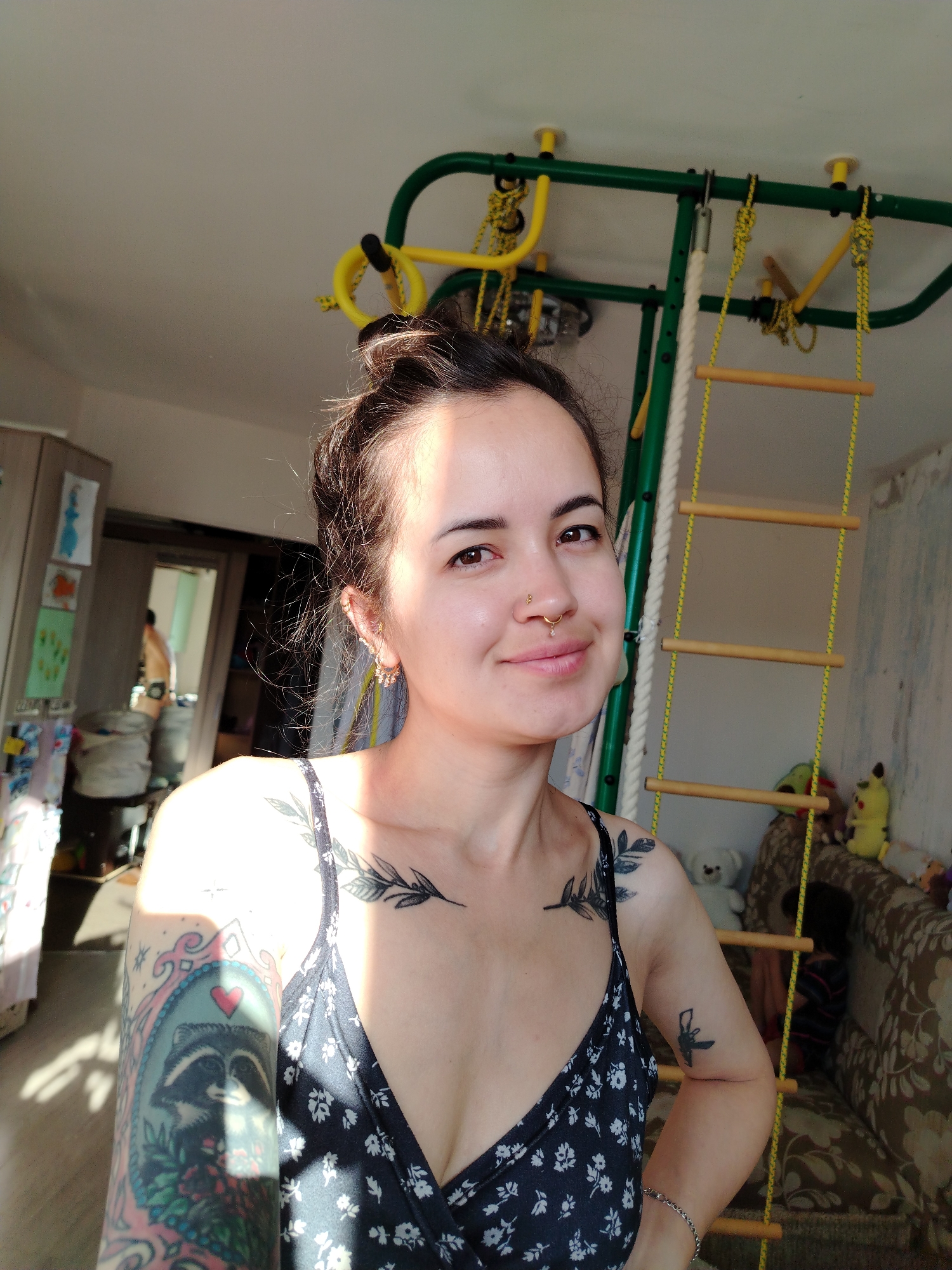 The photograph depicts a young woman with dark hair pulled up in a bun, a nose ring, and extensive tattoos on her arms and chest. She is wearing a dark, floral-patterned dress. She is posing indoors, with a natural light source creating strong shadows on her face and upper body.
In the background, a green and yellow indoor playground structure is visible, along with a ladder made of wooden rungs and yellow rope. To the left, a wooden cabinet with children's drawings attached to it can be seen. A mirror is also present, reflecting a portion of the room, including a person with their back to the camera. The environment appears to be a residential space, possibly a living room or playroom, given the presence of stuffed animals on a patterned sofa.
The photograph was taken in Noviy, Russia.