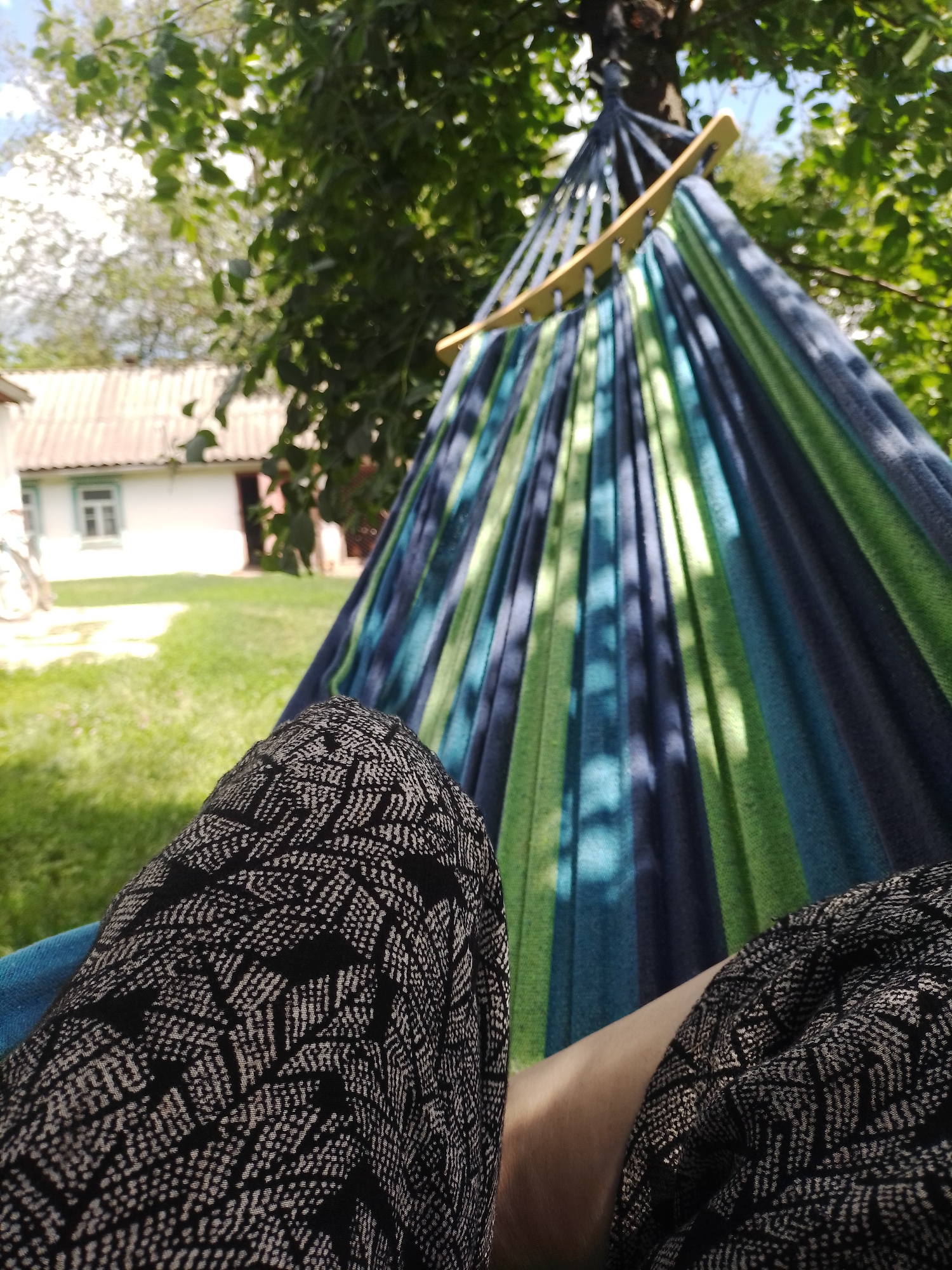 In Ustyvytsia, Ukraine, a first-person perspective shows a person lying in a hammock outdoors. The hammock is made of striped fabric in shades of blue and green, suspended from a tree. The foreground is dominated by the person's legs, covered in black pants with a white geometric pattern. Behind the hammock, a white building with a light-colored roof and a single window is visible in the background, surrounded by green grass and trees. The sky is partially visible through the leaves, with some clouds. The scene suggests a relaxed outdoor setting.