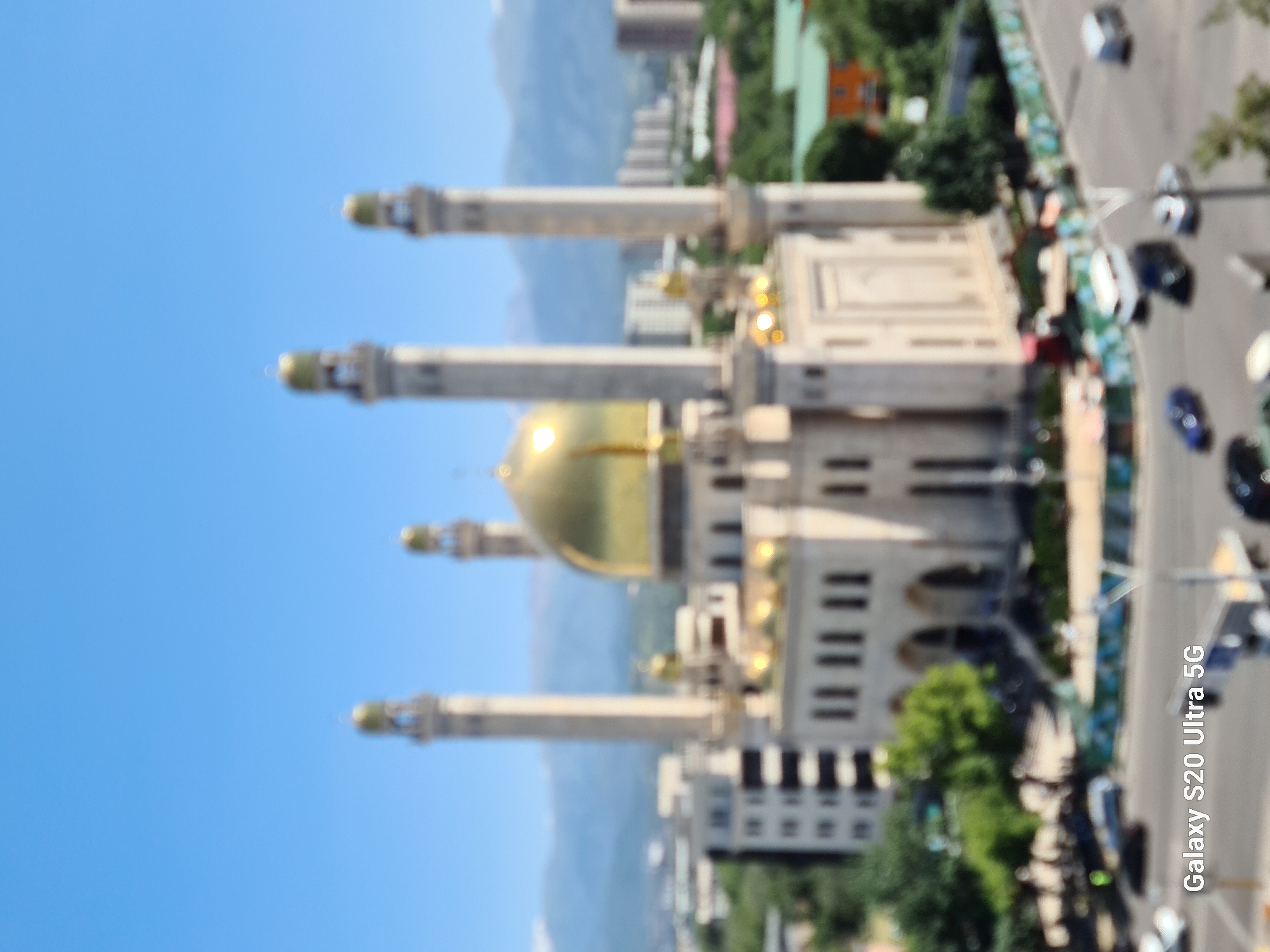 A blurry exterior, daytime shot shows the central mosque of Almaty, Kazakhstan. The mosque is light-colored with a prominent gold dome, flanked by four tall minarets. In the foreground, a street is visible with a few cars driving and parked. The background features buildings and distant mountains under a clear blue sky. The words "Galaxy S20 Ultra 5G" are superimposed on the lower left corner of the image.