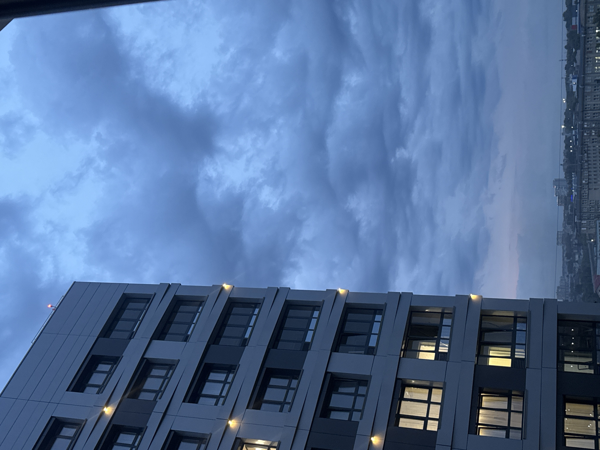 This image depicts a modern apartment building in Novosibirsk, Russia, with a cloudy sky overhead. The building is tall and angular, with multiple floors featuring dark-framed windows and some with lights on inside. The exterior appears to be clad in grey panels. A small red light is visible on the rooftop of the building, likely an aviation warning light. Below the apartment building, a cityscape with several other buildings and structures is visible in the distance, suggesting an urban environment. The sky is filled with heavy, dark clouds, indicating overcast or potentially stormy weather. The overall impression is one of an urban twilight scene.