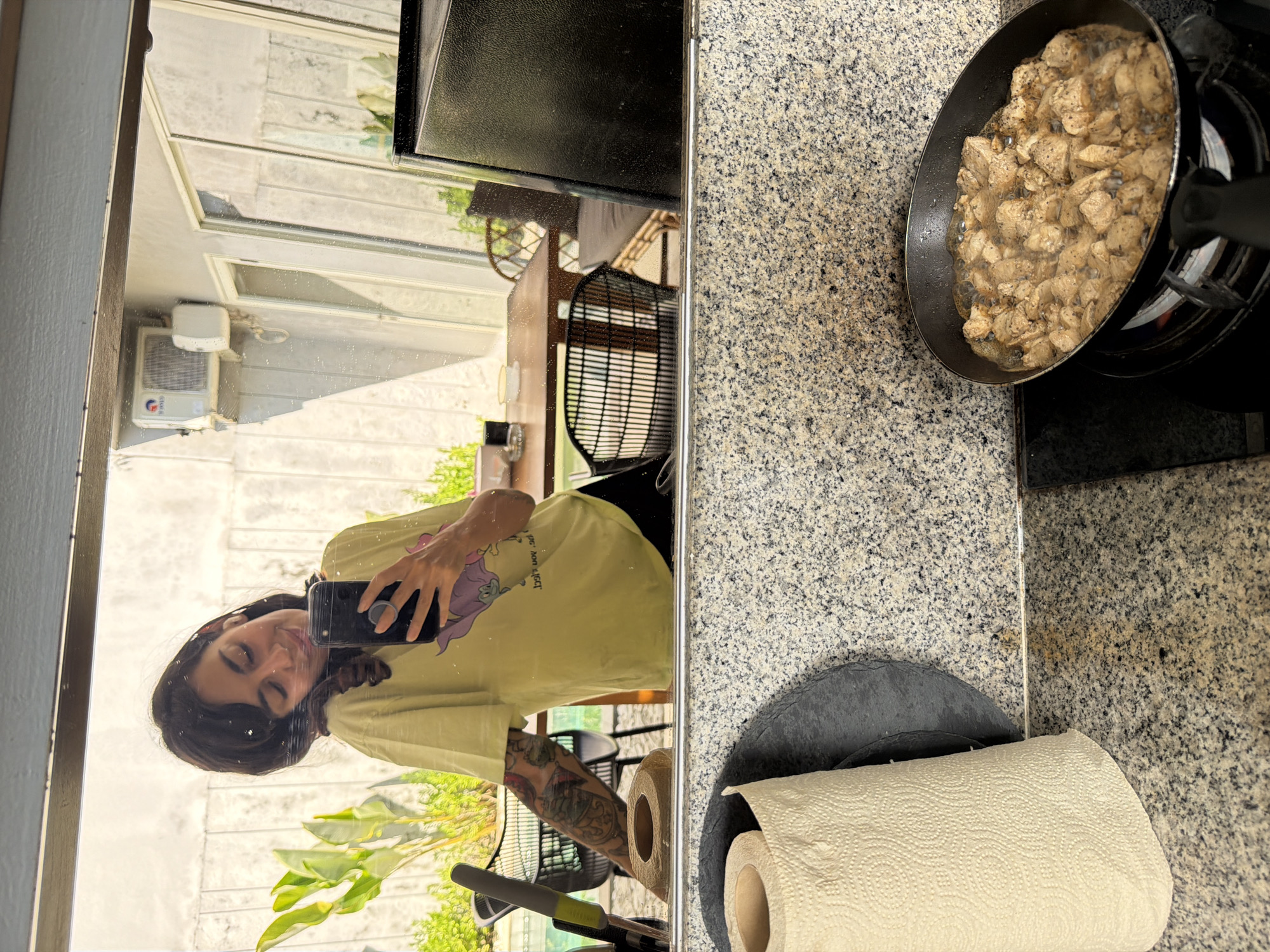 A woman is taking a selfie in a mirror in what appears to be a modern kitchen and outdoor living space. She is wearing a light yellow t-shirt with a cartoon character on it and has tattoos on her arm. She is holding a smartphone with a pop socket. In the foreground, on the kitchen counter, there is a roll of paper towels and a black circular placemat. A black frying pan containing cubed meat is cooking on a black stovetop. In the background, reflected in the mirror, is an outdoor dining area with a wooden table and chairs, lush green plants, an air conditioning unit on a wall, and a swimming pool. The overall impression is a casual and bright domestic setting, likely a home or a villa.