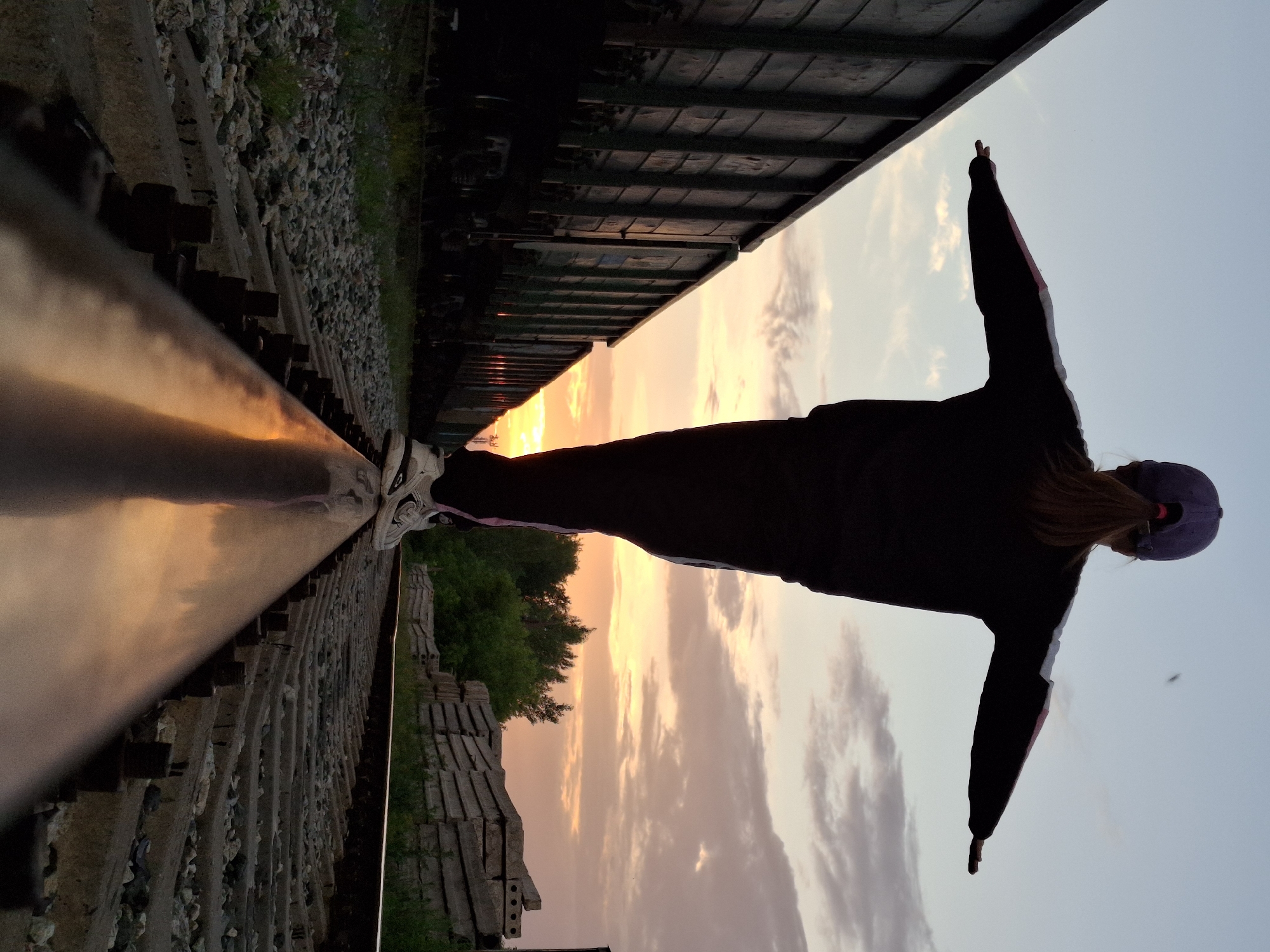 A full-body shot captures the back of a person, silhouetted against a dusky sky as they balance on a railway track. The individual, wearing a purple baseball cap and a dark tracksuit with white and pink stripes on the sleeves, has their arms extended to the sides for balance. Their hair, tied in a ponytail, falls down their back. They are wearing white sneakers with black accents. The setting appears to be a railway yard, with multiple train cars visible to the left of the frame, their dark, weathered surfaces contrasting with the warm hues of the sunset. To the right, a pile of concrete blocks and green foliage are present. The railway tracks themselves are the central focus, with the person positioned on one of the rails. The ground surrounding the tracks is covered with gravel and sparse vegetation. The sky above transitions from a pale blue near the top to shades of orange and pink near the horizon, indicating either sunrise or sunset. The reflection of the sunset is visible on the curved surface of the railway track beneath the person's feet. This scene is located in the city of Chaikovsky, Russia.