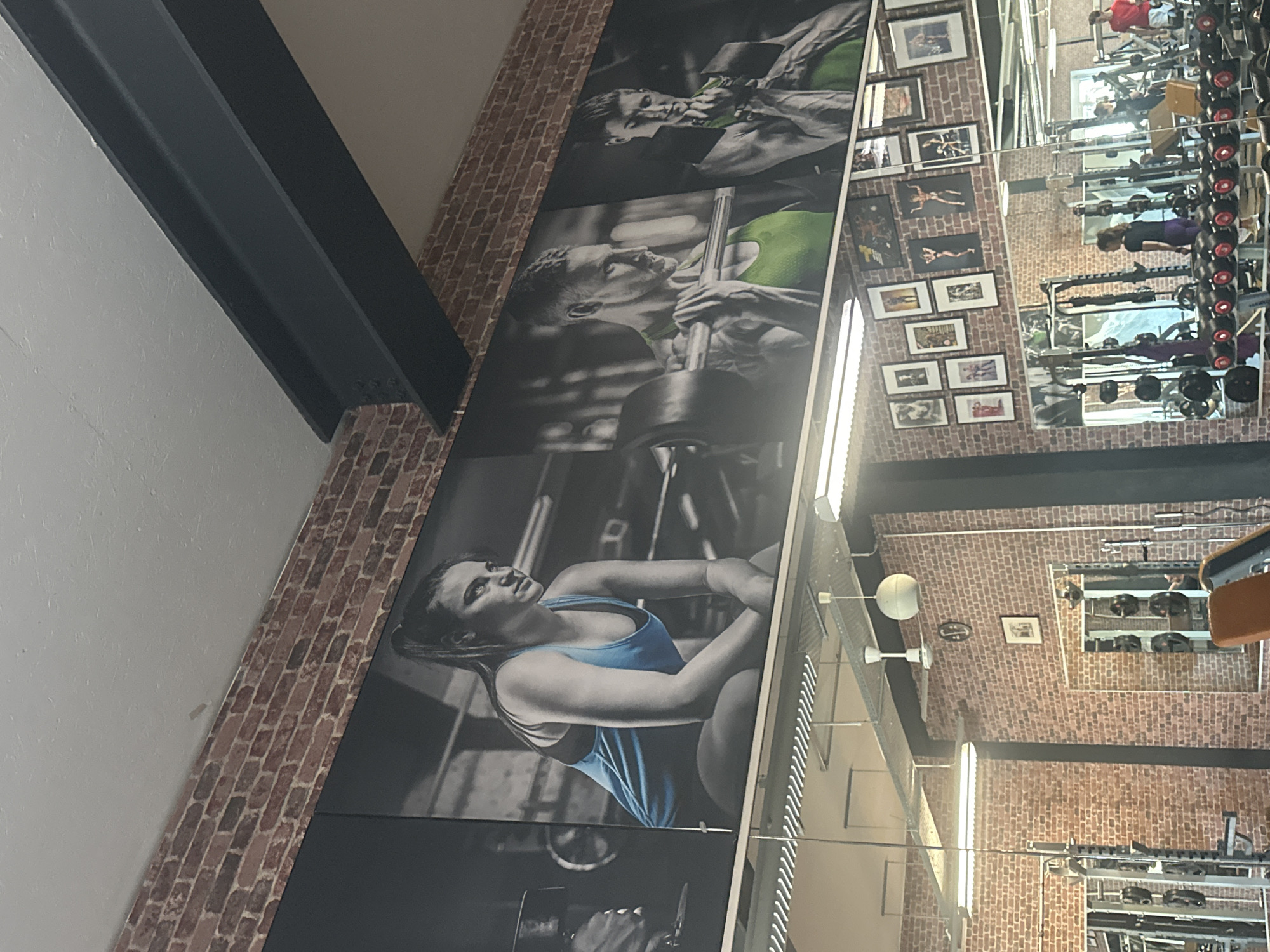 The image shows a gym interior, likely in Brunnthal, Germany. The walls are a mix of brick and painted surfaces. A large mural depicts people lifting weights, suggesting a fitness-oriented environment. Reflections in the mirrors reveal gym equipment, including weights and machines. There are also smaller framed pictures on the walls. The structural elements of the building, such as support beams, are visible.