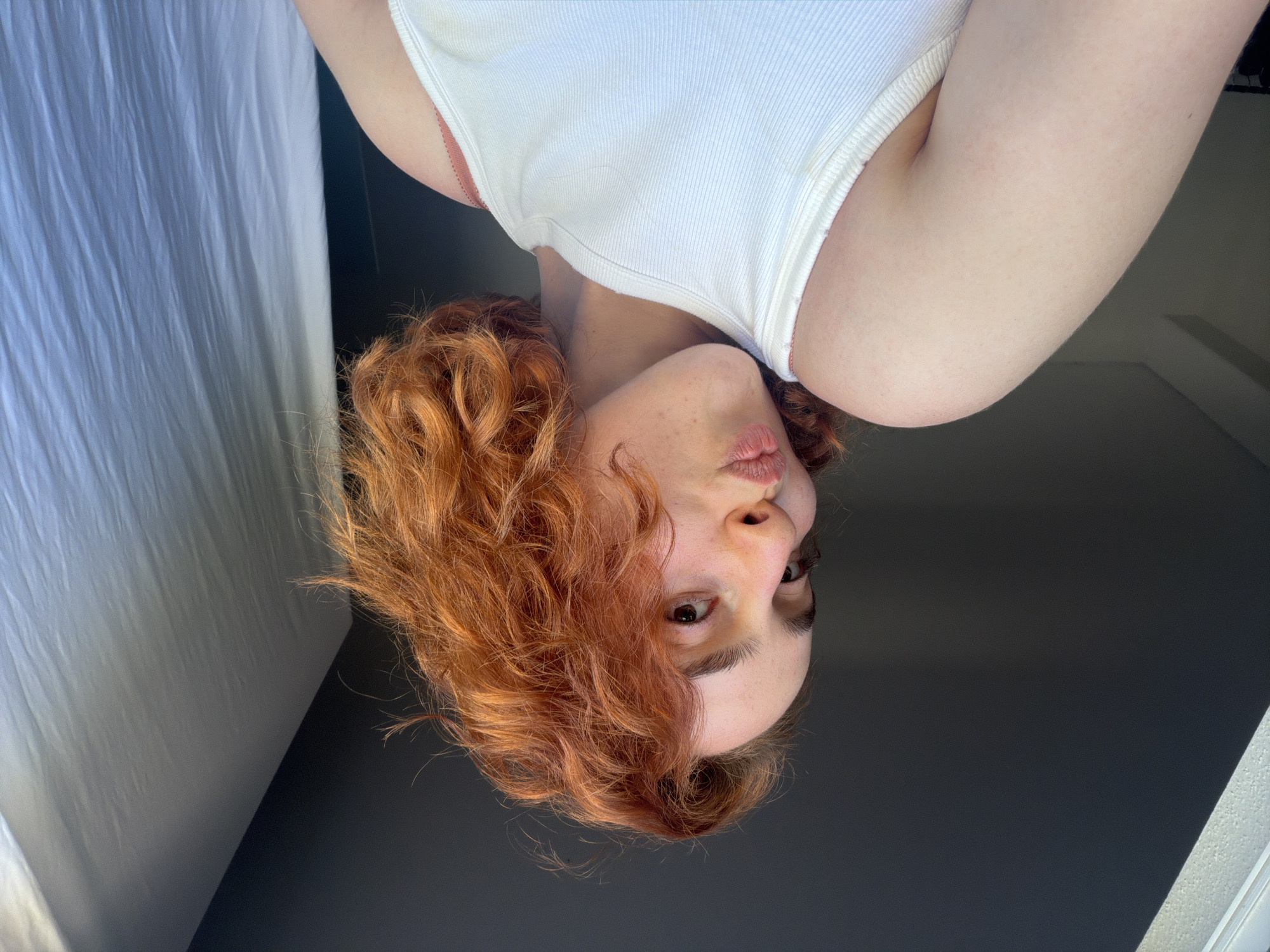 An individual with ginger hair and fair skin is posing for a selfie in what appears to be a room. The individual is wearing a white ribbed tank top. The background suggests an indoor setting with natural light coming from the left, illuminating a white curtain on the right. The lighting creates a contrast between the shadowed left side of the image and the brighter right side. There are no discernible landmarks or context that would suggest a specific location within Strasbourg, France.