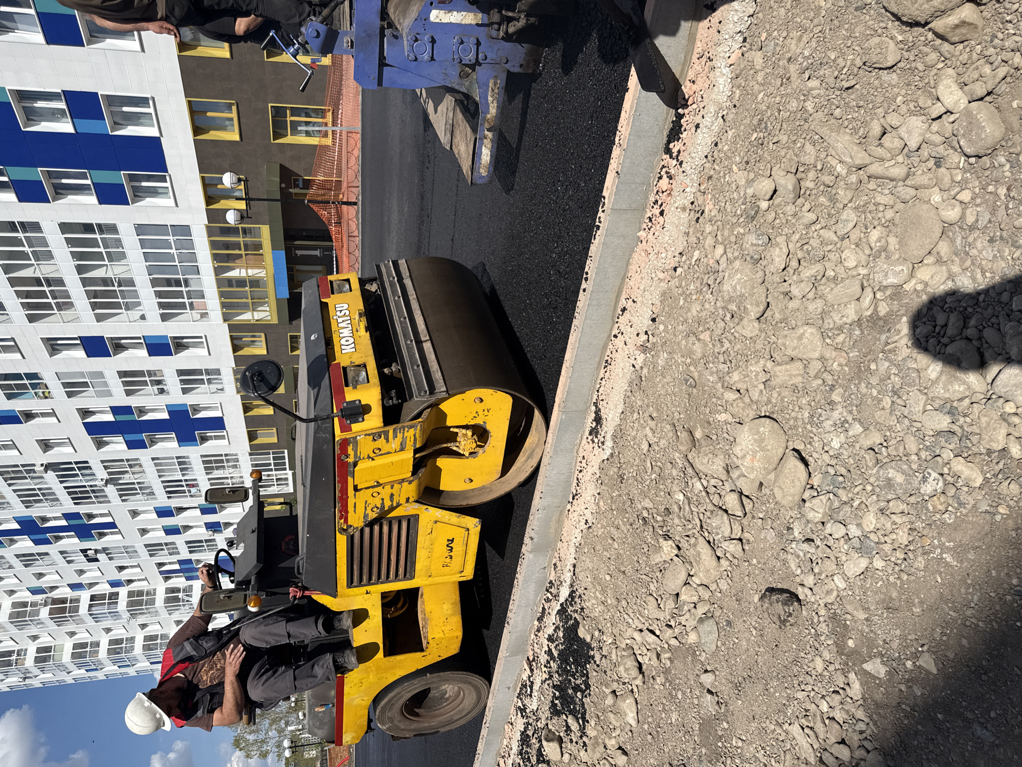 A street resurfacing project is underway. A male worker in a hardhat operates a Komatsu asphalt roller on a newly paved, black asphalt surface bordered by a concrete curb and a gravel shoulder. A second, unidentified worker stands at the edge of the pavement. In the background is a multi-story apartment building, located in the city of Krasnoyarsk, Russia.