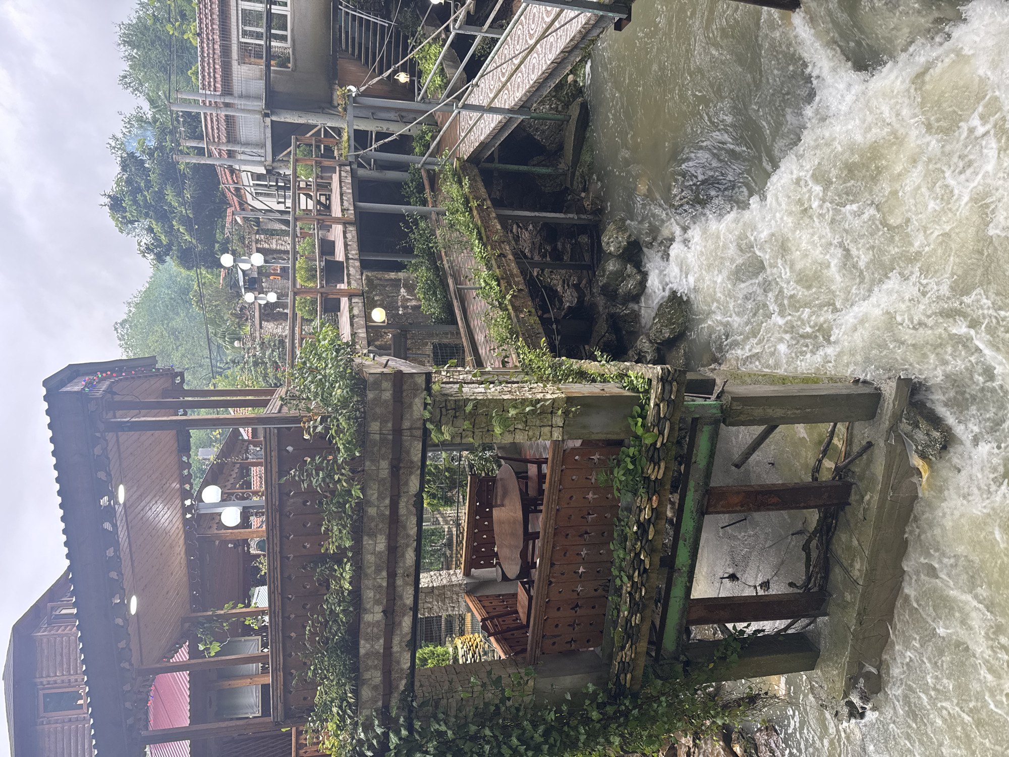 The image captures a scene in what appears to be Tsabal, Abkhazia, featuring a building complex alongside a rapidly flowing river or stream.
The primary subjects are the buildings constructed along the waterway, which seem to be multiple stories high and built with a mix of stone, brick, and wood materials. Sections of the structure extend over the water on support pillars. There is vegetation, including vines, growing on the walls and supports of the building.
The river is a prominent element, with turbulent water flowing swiftly and foaming. The buildings seem to be positioned to take advantage of the water feature.
A pedestrian bridge is located higher above the river, connecting two sections of the buildings.
The lighting is diffused, suggesting an overcast day. The overall environment gives the impression of a tourist location.