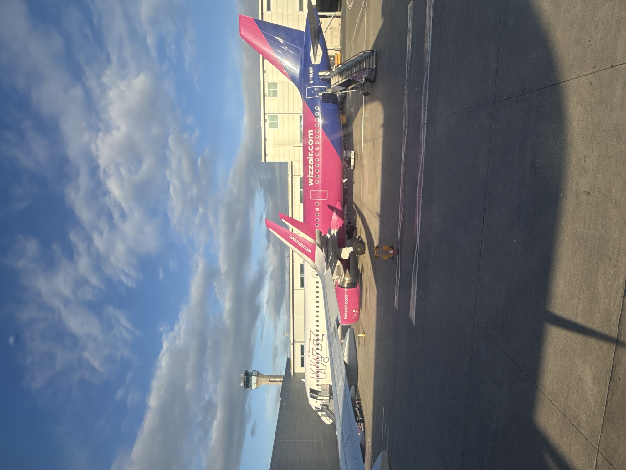 Two Wizz Air airplanes are parked on the tarmac at an airport. The airplane in the foreground is primarily pink and blue with "wizzair.com" written on its side. A set of stairs is extended to the rear passenger door. The airplane behind it is predominantly white with "Wizz Air" branding visible on its fuselage. In the background, a tall control tower stands against a sky with scattered clouds. The ground is asphalt with markings.