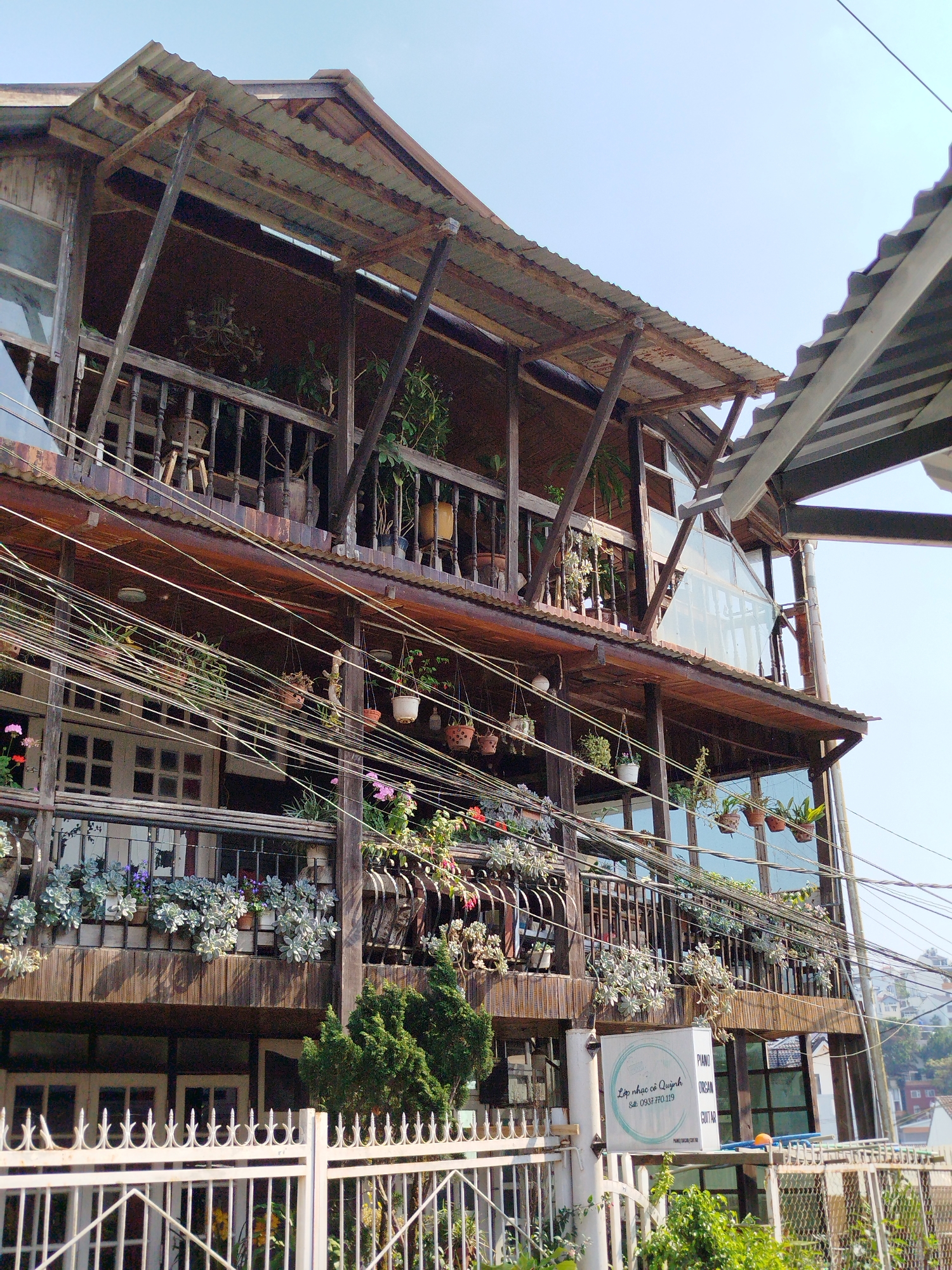 The image shows a multi-story wooden building with balconies, located in Da Lat, Vietnam. The building has a rustic appearance with exposed wooden beams and a corrugated metal roof. Potted plants and flowers adorn the balconies, creating a vibrant display. Power lines crisscross in front of the building. A white fence runs along the base of the building, and a sign with Vietnamese text and the words "Piano, Organ, Guitar" is visible. The sky is clear and light blue.