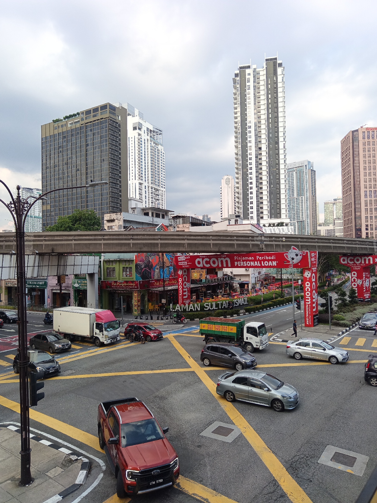 An aerial, eye-level view captures a busy intersection in Kuala Lumpur, Malaysia, under a cloudy sky. The scene is dominated by urban architecture, including several tall, modern skyscrapers. In the foreground, a red Ford Ranger pickup truck with license plate "UTM 5738" is stopped at a traffic light. Yellow road markings delineate the intersection, and pedestrian crossings are visible. Several other vehicles, including cars and a white box truck, are present on the road. Overhead, a monorail track runs across the scene, supported by concrete pillars. On the ground level, storefronts with colorful signage line the street. Red banners with the word "acom" and "PERSONAL LOAN" are prominently displayed, advertising financial services. The environment appears to be a commercial district with a mix of contemporary and older buildings.