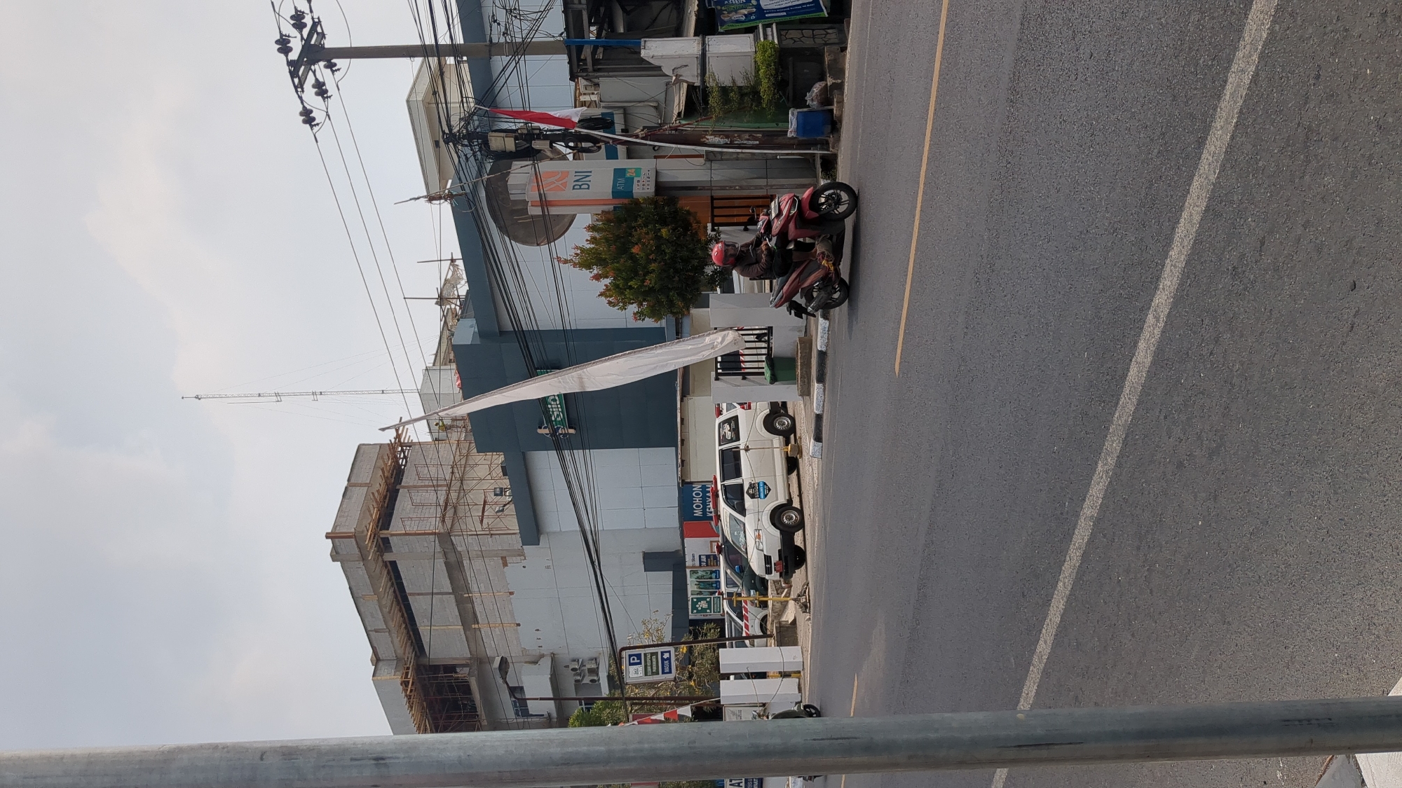 The image depicts a street scene in Purwakarta, Indonesia. In the foreground, a dark asphalt road is visible with white and yellow lane markings. A gray metal pole stands to the left, partially obscuring the view. In the middle ground, a white van with red flashing lights is parked, displaying text that reads "MOHON KEMBALI JALAN". To the right of the van, a person on a red motorcycle, wearing a red helmet, is riding on the road. Behind the parked van, a building with the BNI logo and an ATM 24 sign is visible. Further in the background, a large building under construction is present, with scaffolding and construction materials. The sky is overcast.