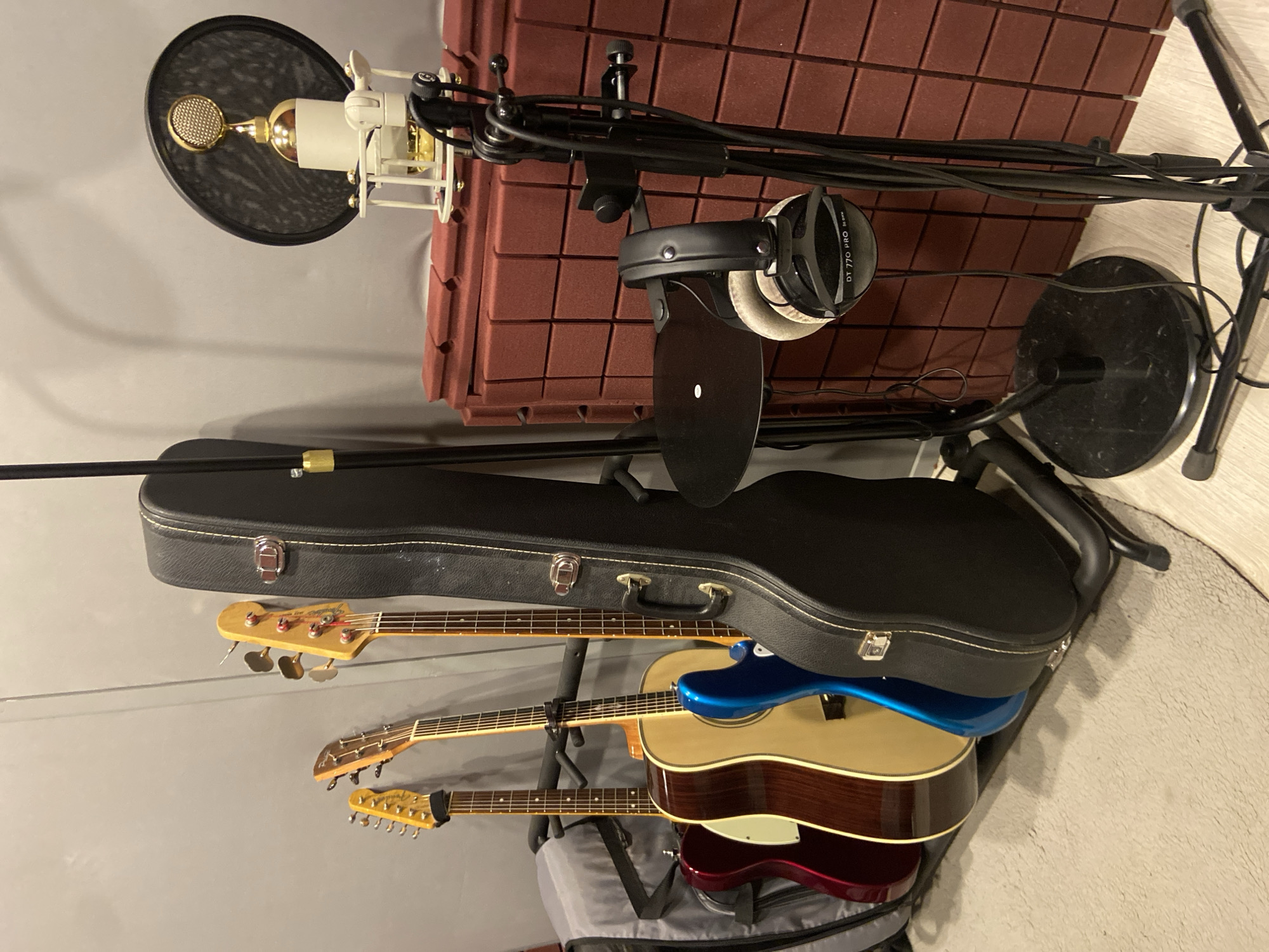 This image, stated to be from Kazan, Russia, depicts a music recording setup within what appears to be a dedicated studio space or music corner. On the left, a multi-guitar stand holds four instruments: a red electric guitar resembling a Fender Telecaster, an acoustic guitar with a natural wood top and dark back/sides (featuring a capo on its neck), a blue electric guitar, and a bass guitar. A dark grey soft gig bag is partially visible behind the guitars, and a black hard-shell guitar case stands upright to the right of the bass. The guitars rest on a light grey rug covering a light-colored wooden floor. The wall behind the guitars is a plain grey.
To the right, a studio microphone setup is positioned against a wall covered with reddish-brown acoustic foam panels arranged in a grid. A golden-colored large-diaphragm condenser microphone is held within a white shock mount, with a black pop filter (featuring a gold rim) positioned directly in front of it. This microphone assembly is mounted on a robust black microphone stand with a circular base. Adjacent to it, a separate black stand features a small circular shelf and a headphone holder from which a pair of black Beyerdynamic DT 770 PRO 80 OHM over-ear headphones hangs. Wires and cables are visible around the microphone and headphone stands. No individuals are present in the scene.