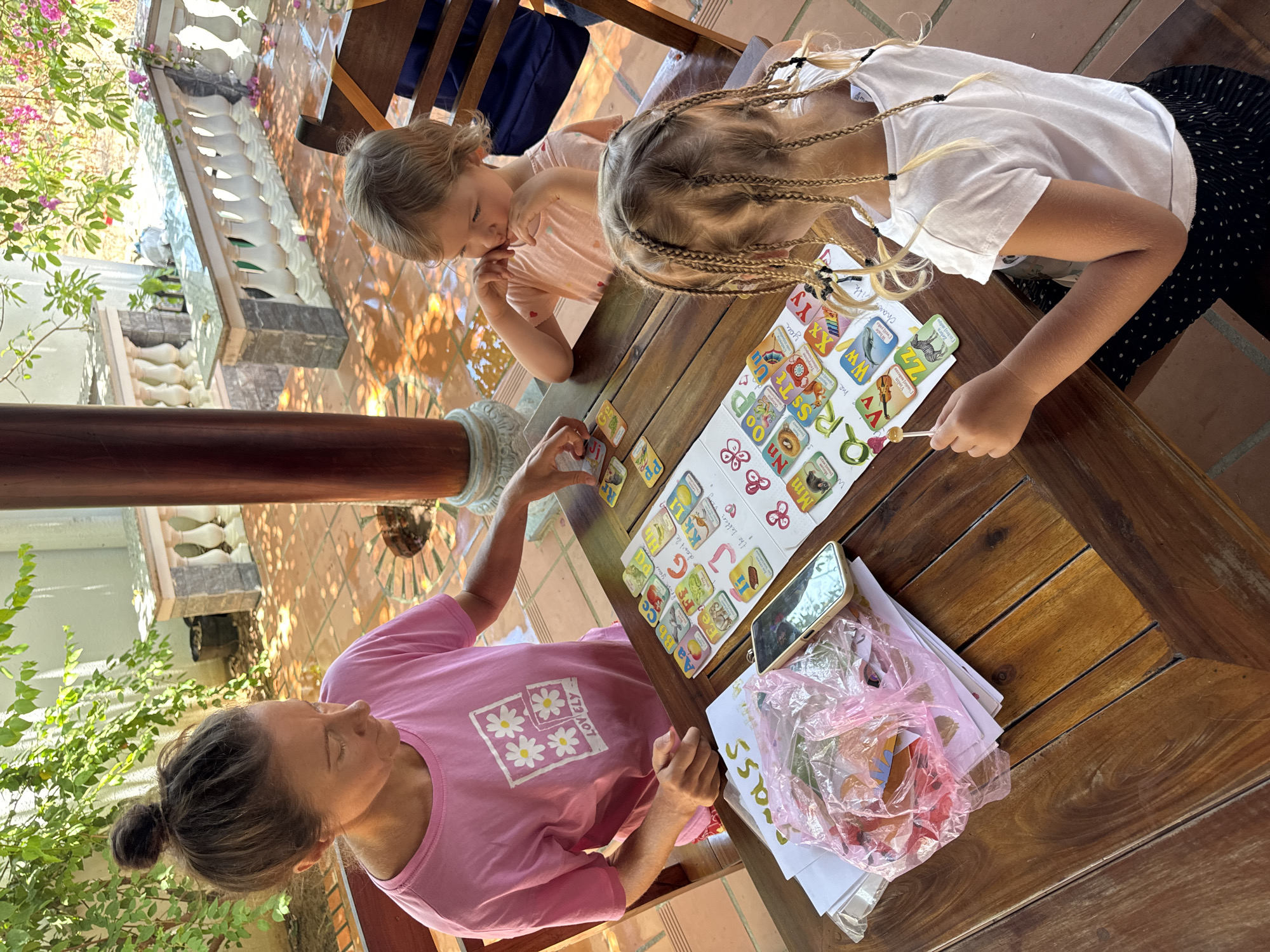 The image captures a moment of interaction and education at a table in what seems to be an open or semi-open space, possibly a patio or veranda in Mũi Né, Vietnam. A woman wearing a pink shirt with the word "LOVELY" on it sits at the table, engaged in a learning activity with two young girls. The girls, one with braided hair and the other with shorter hair, are focused on an alphabet-themed learning game laid out on the wooden table.
A phone is placed among the learning materials and some plastic bags filled with unidentified objects. The environment features tiles on the floor and sunlight filtering through foliage, suggesting a tropical setting. The surrounding architecture includes detailed balustrades, indicative of local building styles. The image composition emphasizes the shared learning experience and the intimate family setting.