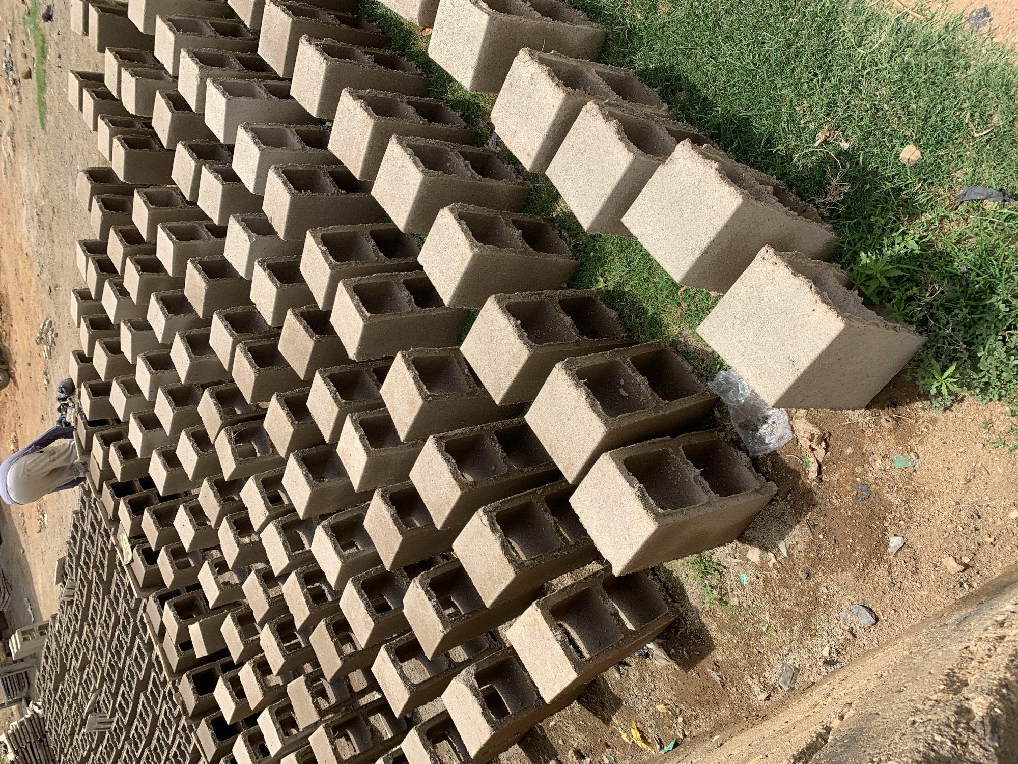 The scene is located in Rafin Par, Nigeria. It depicts a large number of hollow concrete blocks, likely freshly made, arranged in neat rows. The blocks are light gray in color and feature two cylindrical voids running vertically. Some blocks are placed on grass, while others sit on bare ground. A person wearing a white head covering and light-colored clothing is visible near the blocks on the left side of the frame. Debris, including plastic and glass shards, is scattered on the ground. The environment is sunny.
