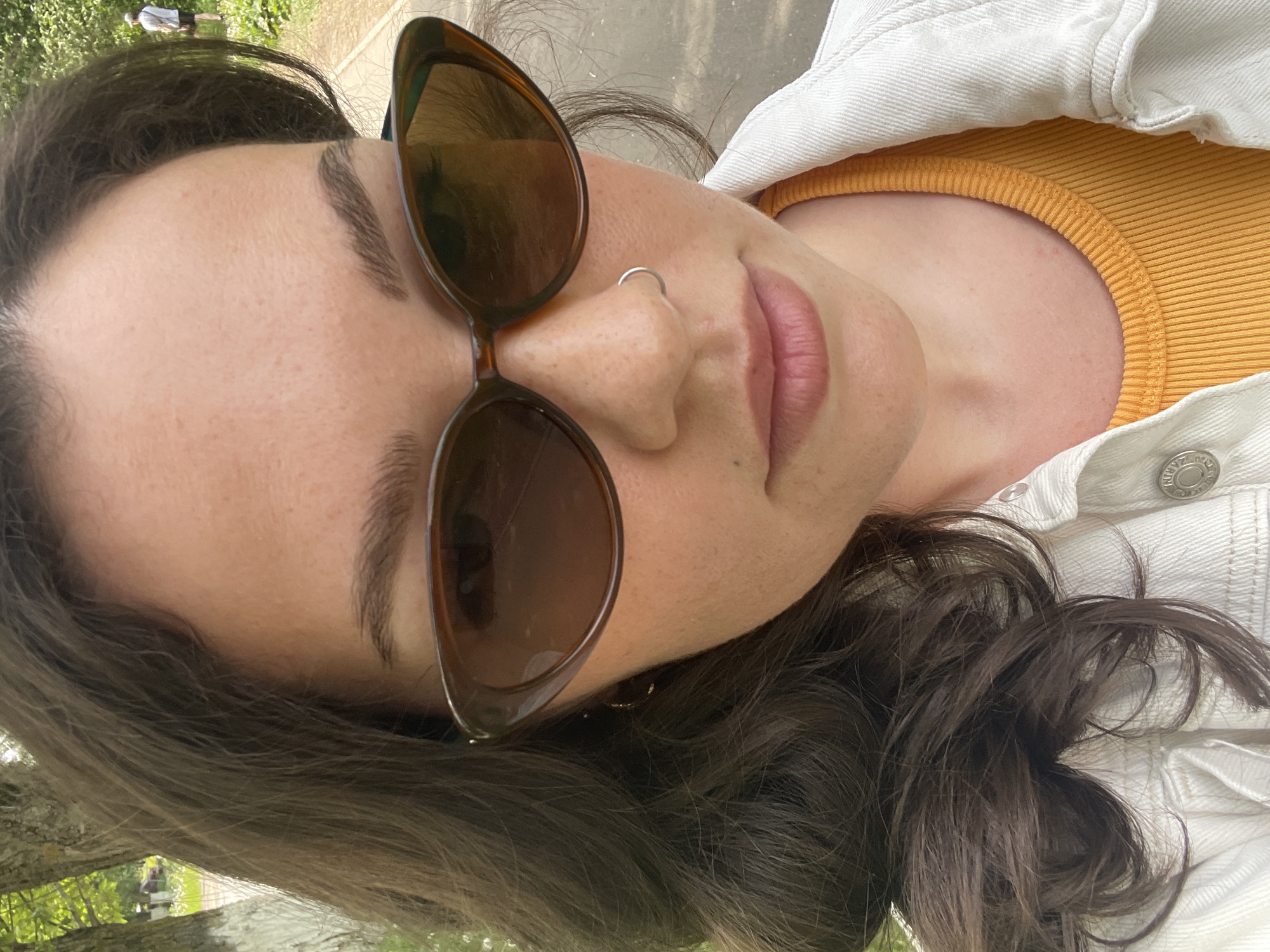 A close-up, rotated selfie of a woman outdoors, likely in a park. She has dark brown hair, fair skin, and is wearing brown sunglasses with a round frame. She has a nose ring and is wearing a yellow top with a white denim jacket over it. In the background, blurred, are trees and a path, with a person walking on the path in the distance. The location is not able to be determined as Izhevsk, Russia.
