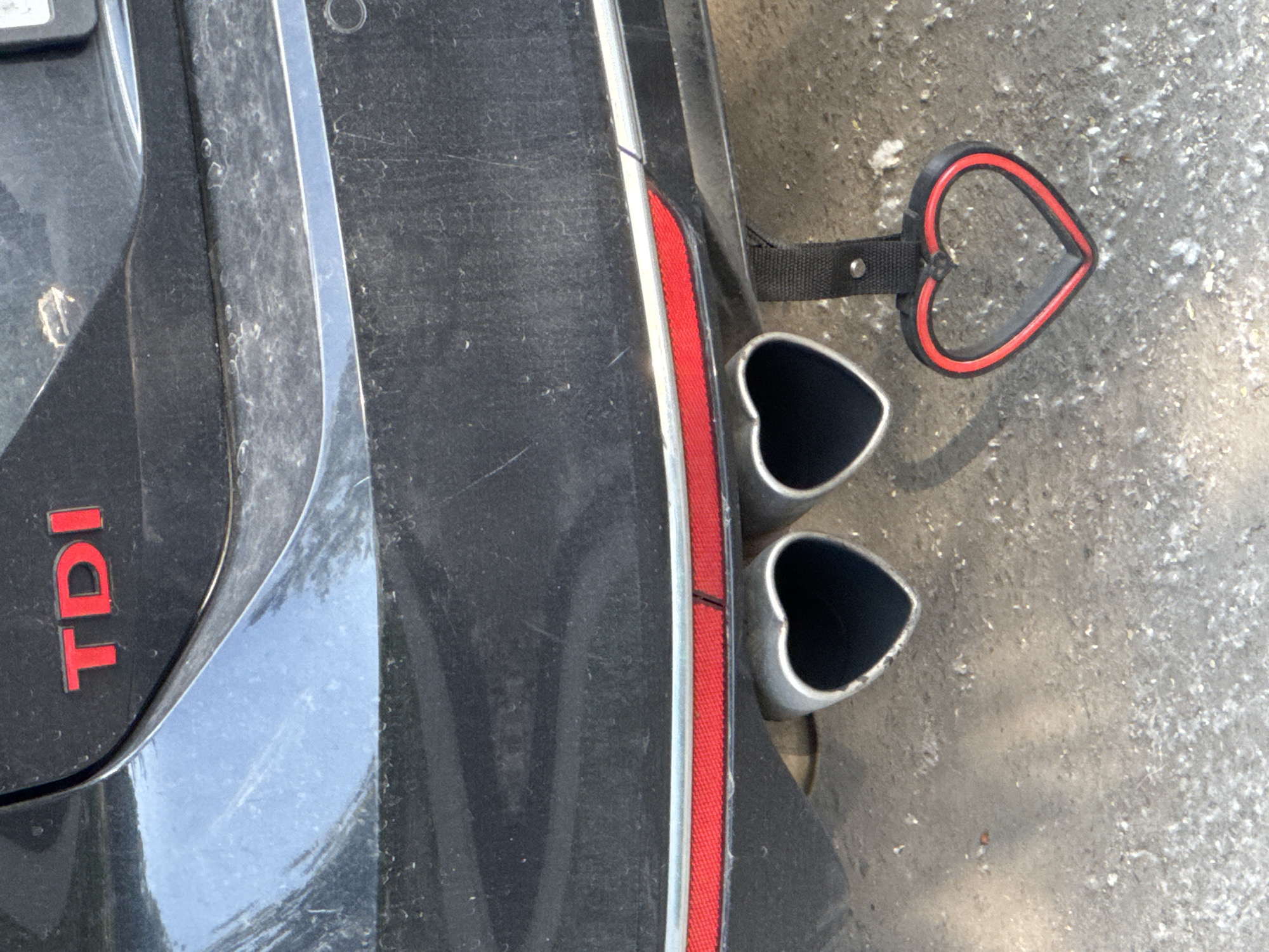 The media file displays a close-up, high-angle shot of the rear lower section of a dark-colored, possibly black, car parked outdoors in what appears to be Moscow, Russia. The car's surface is noticeably dusty and shows some minor wear.
The primary subjects are the car's bumper area and its custom exhaust system. Two heart-shaped exhaust pipe tips, made of silver-toned metal, protrude from beneath the bumper. Immediately to the right of these exhaust tips, a decorative, heart-shaped object is visible. This object is red and black, appearing to be a flexible material, and is attached by a black fabric strap to what might be a tow hook or part of the car's undercarriage.
On the left side of the image, the red letters "TDI" are prominently displayed on the car's body, indicating the engine type. A red reflective strip or trim is integrated into the car's design between the main body and the lower bumper section, running vertically. The ground beneath the car is a rough, dusty asphalt or concrete surface.
The lighting suggests it is daytime, and the dry, dusty conditions indicate fair weather. The heart-shaped elements, both the exhaust tips and the hanging decoration, suggest a personalized and distinctive aesthetic chosen by the car owner. No human subjects or active events are depicted; the image is a static snapshot focusing on the vehicle's rear detailing.