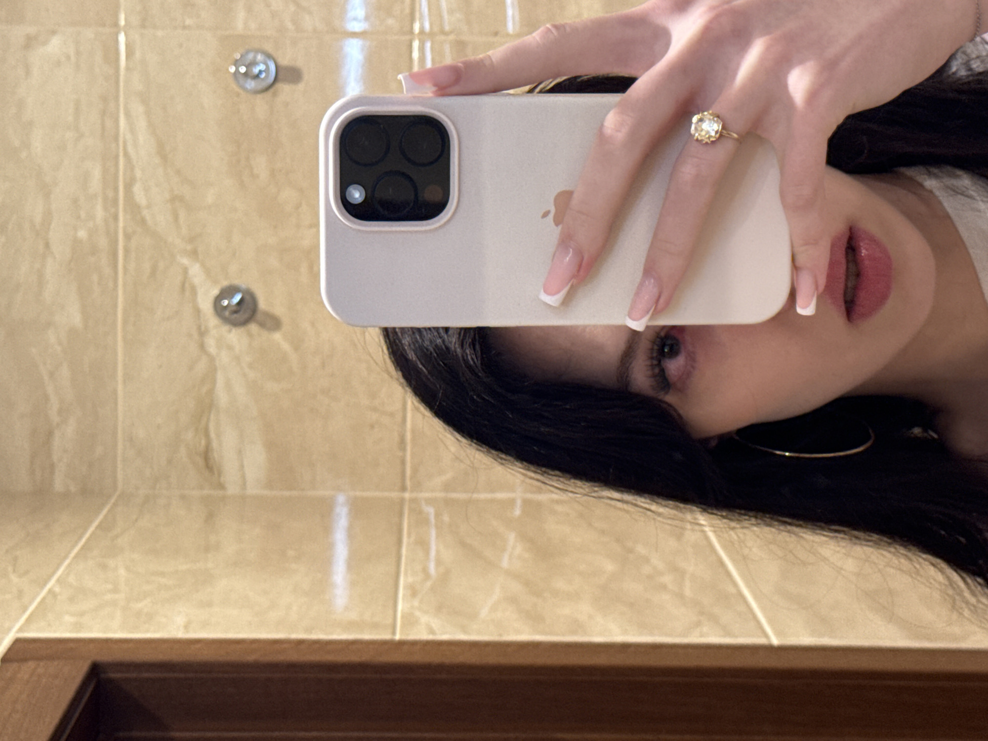 A young woman is taking a selfie in a bathroom. She is holding a white iPhone in her right hand, and the phone is angled to capture her reflection in a mirror. Her left eye and part of her forehead are visible, along with her dark hair and a large hoop earring on her left ear. She is wearing a light-colored top. Her right hand, adorned with a flower-shaped ring and French manicured nails, is resting on the back of the phone, partially obscuring her face. The background appears to be a tiled wall with a wooden door frame on the left. The scene suggests a private residential setting, likely a bathroom.