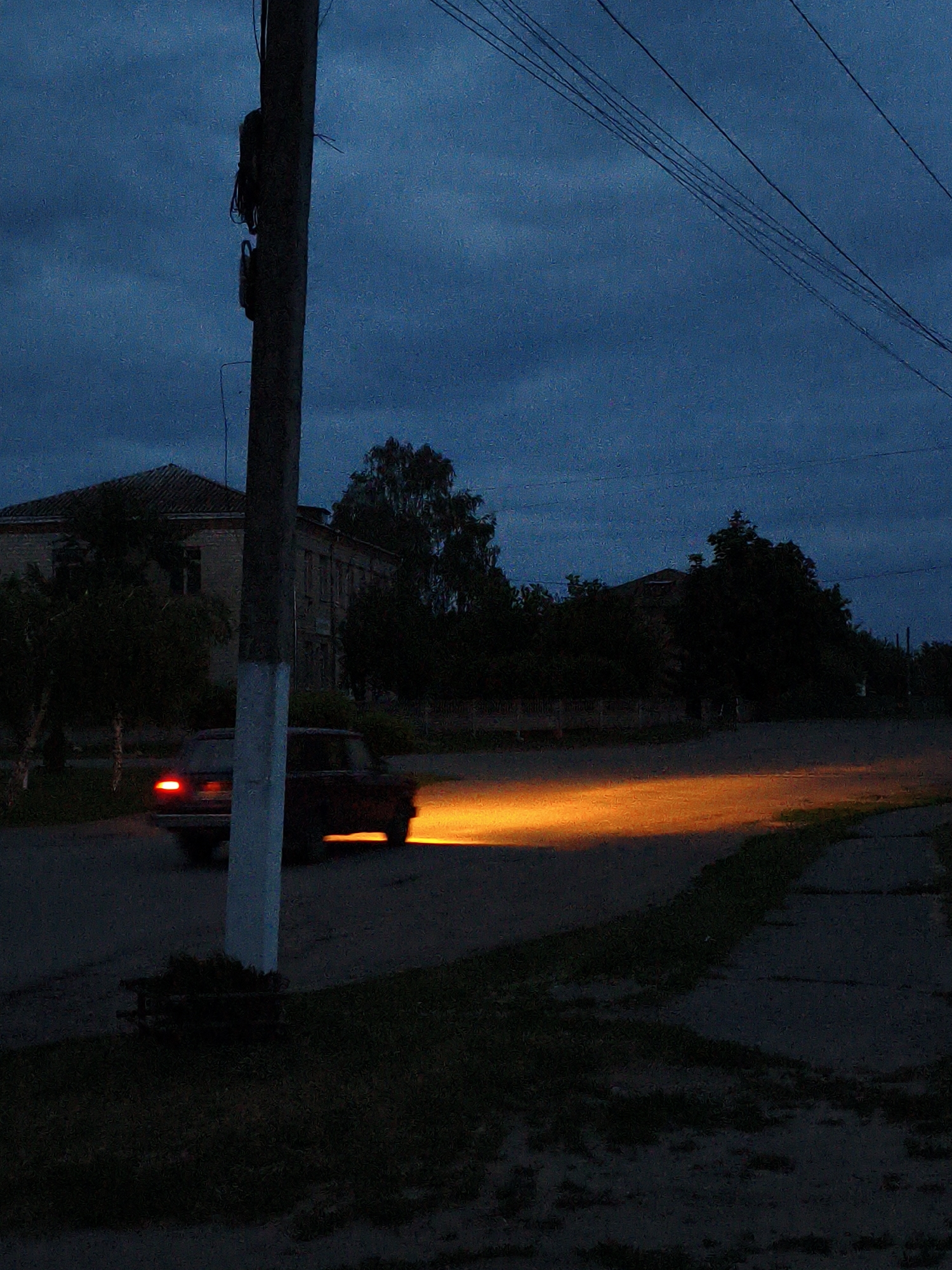 This nighttime image depicts a quiet street scene in Ustyvytsia, Ukraine, under a dark, cloudy blue sky. The central subject is a car, possibly an older model, moving from left to right across the frame. Its headlights cast a vivid, elongated orange-yellow beam onto the unpaved road due to motion blur, highlighting its movement, while a red taillight is also visible.
In the foreground, a tall utility pole, painted white near its base, stands prominently on the left, with multiple power lines stretching diagonally across the upper portion of the sky. To the left of the pole, a dark, silhouetted building with a tiled roof is partially visible, surrounded by trees. More dark trees and distant structures line the street in the background. The road appears to be unpaved or poorly maintained, with grassy verges. A small, low, dark structure is situated at the base of the utility pole. The overall impression is one of a quiet, rural or semi-rural setting illuminated only by the moving car's lights. There is no visible text in the image.