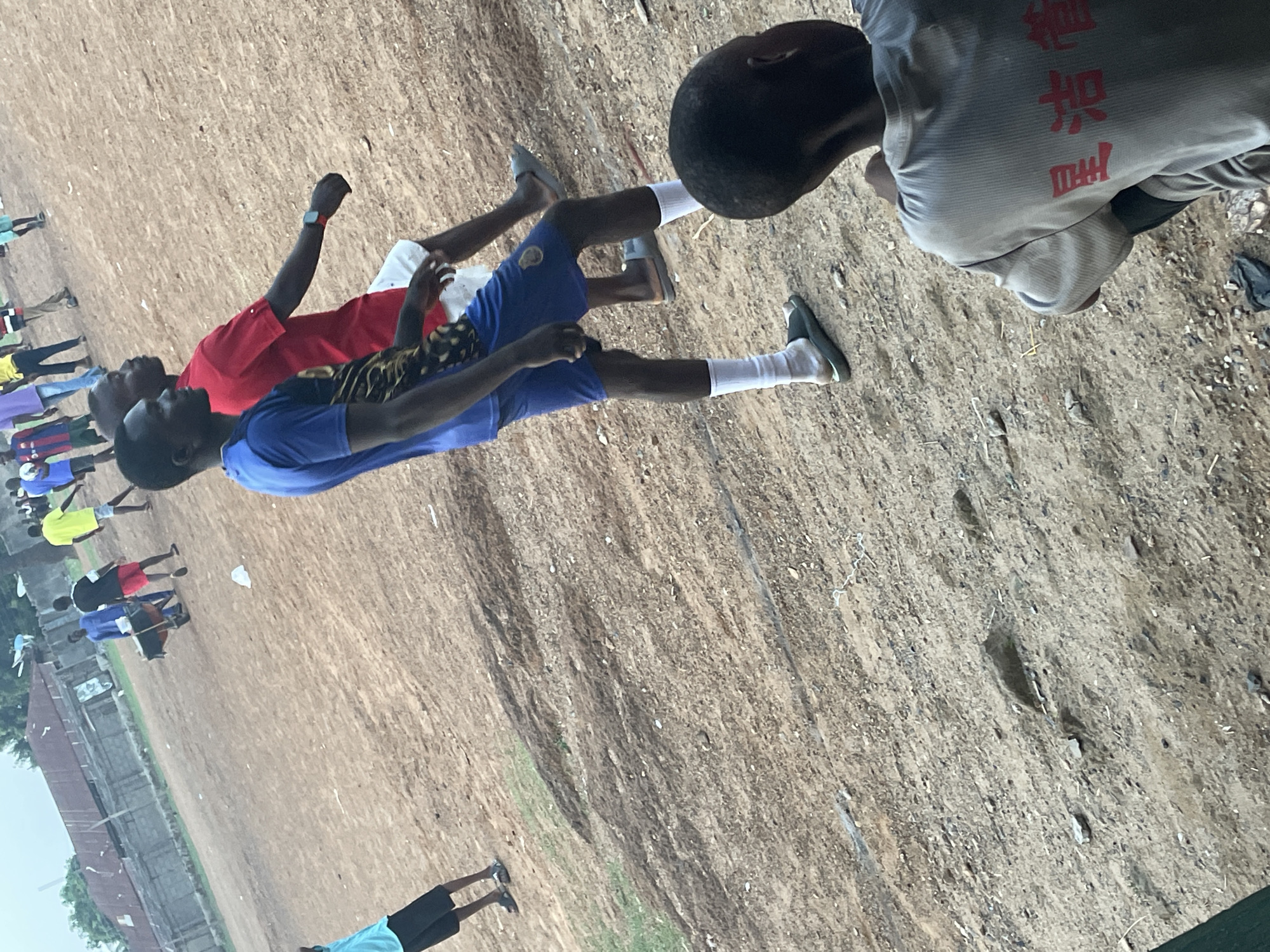 The image shows a group of young men and boys in Akwanga, Nigeria, standing on a brown dirt field. Some are wearing athletic attire, including red, blue, and yellow shirts, white socks, and shorts. Some individuals are lined up as if for an activity. A building is visible in the background.