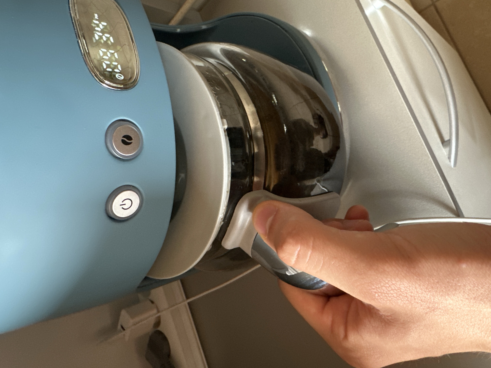 An overhead shot shows a hand holding the carafe of a light blue coffee maker. The coffee maker has a digital clock displaying "09:34 PM" and two buttons. The carafe is glass and filled with a dark liquid. The coffee maker sits on a white countertop with a drawer below. The background is blurry, but appears to be a kitchen environment with a light-colored wall and electrical outlets. The image is not identifiable as being in Anduze, France.