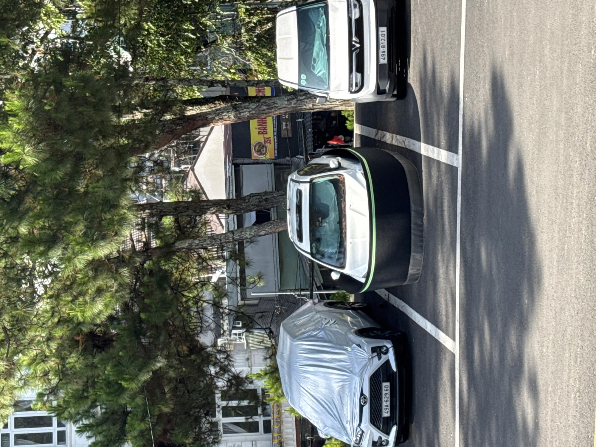 This aerial shot features a street scene, likely in Da Lat, Vietnam, given the license plates on the vehicles. Three vehicles are parked alongside a street with clear parking lines. A vehicle covered in a silver tarp is parked near the bottom, facing the viewer. Slightly above, a white car with a black protective cover is parked. Further up the image, a white van is parked parallel to the other vehicles. Trees flank the left side, partially obscuring buildings in the background. A sign in Vietnamese is visible behind the van. The street surface is asphalt. The overall daylight condition appears bright. No people are directly visible in the frame.