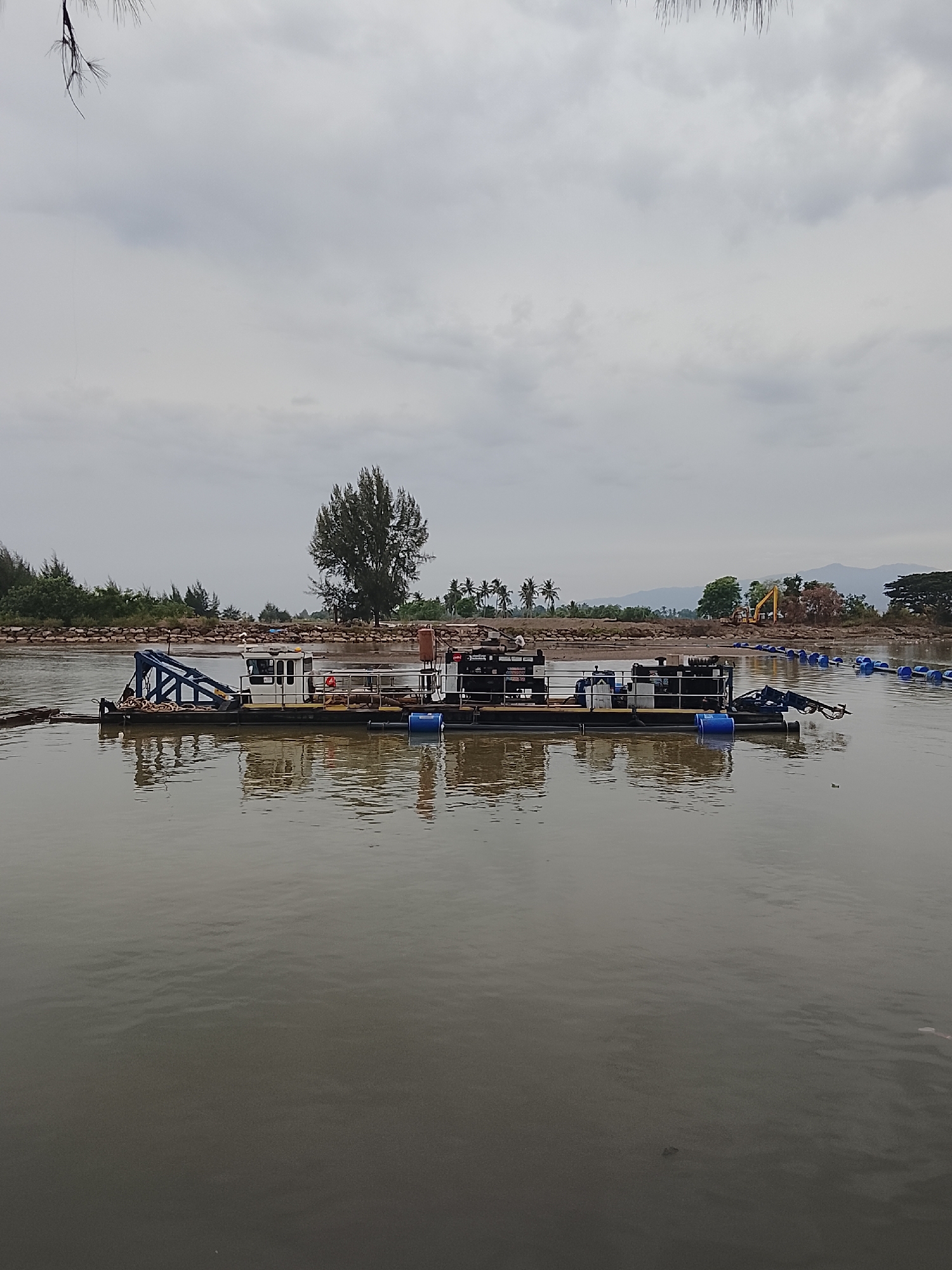 In the city of Reudeuep, Indonesia, a dredging barge is situated on a calm body of water. The barge, predominantly dark in color with white and blue accents, features a prominent blue crane structure on its left side. Several blue barrels are visible on the deck of the barge, along with ropes and other equipment. A long line of connected blue buoys extends from the right side of the barge into the water. The background depicts a tree-lined shore with a rocky embankment. Further inland, construction equipment, including a yellow excavator, is visible, suggesting ongoing development or land reclamation. In the distance, mountains can be seen under a cloudy sky. The water's surface reflects the overcast sky and the objects on the barge.