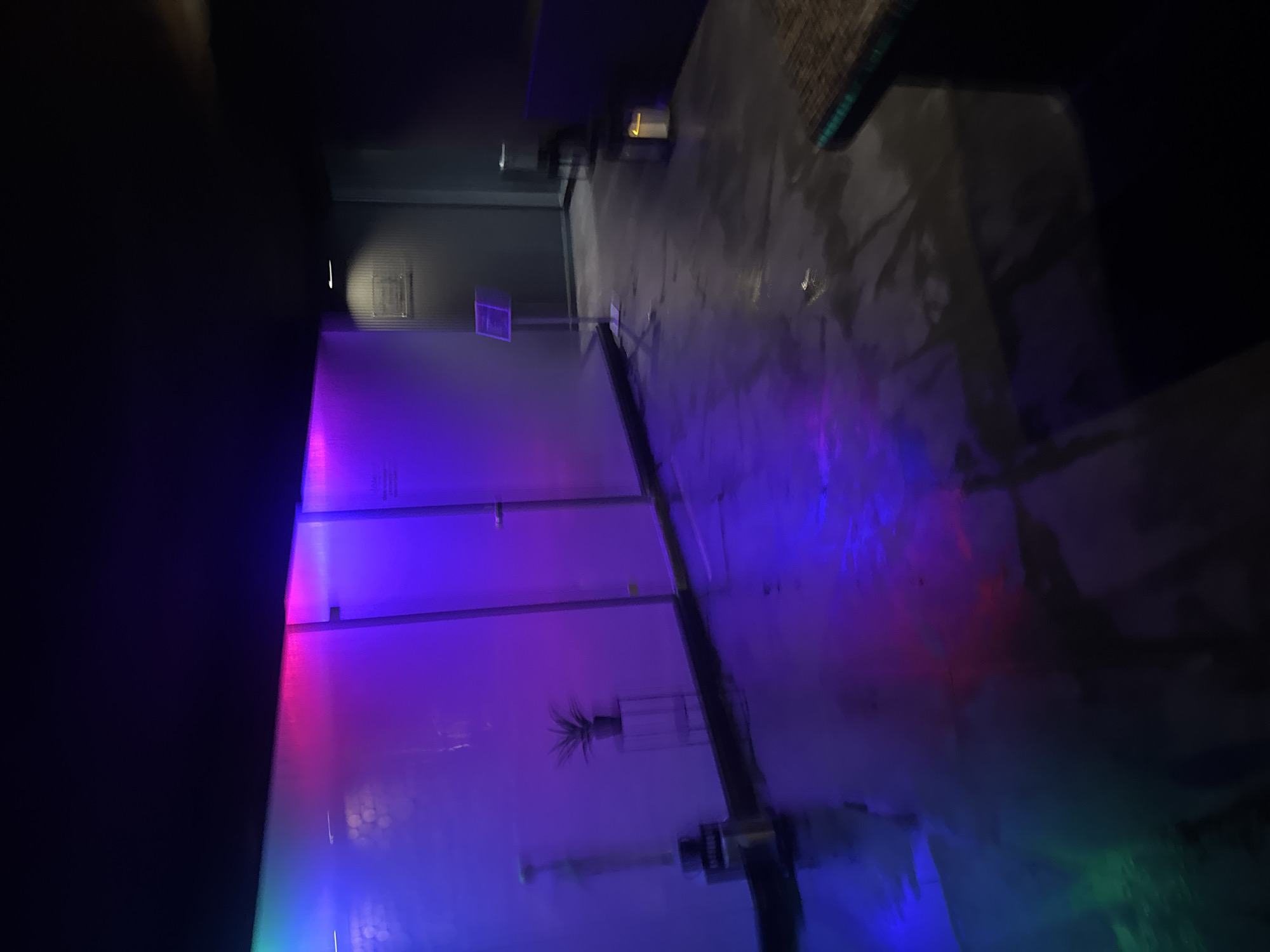 This image captures an interior space located in Plovdiv, Bulgaria, characterized by atmospheric, multicolored LED lighting. The scene presents a modern hallway or lounge area, devoid of any individuals. On the left, a translucent wall is illuminated with a gradient of blue, green, pink, and purple light, which reflects upon the dark, reflective floor, likely concrete or a similar polished material. Against this illuminated wall, a cylindrical metal object, possibly a waste receptacle, is positioned on the far left. Further right, a potted plant stands on a transparent, minimalist support. The wall exhibits vertical divisions, suggesting glass partitions. On the right side, a darker, possibly gray wall features a mounted rectangular sign and, below it, a silver display stand holding a clear information frame. Deeper into the background, an indistinct dark structural element is visible. In the lower right foreground, a dark, textured surface, potentially a table edge or counter, is partially visible, next to which a small, glowing lantern-style lamp rests on the floor. The overhead ceiling area is dark and largely undiscernible.
