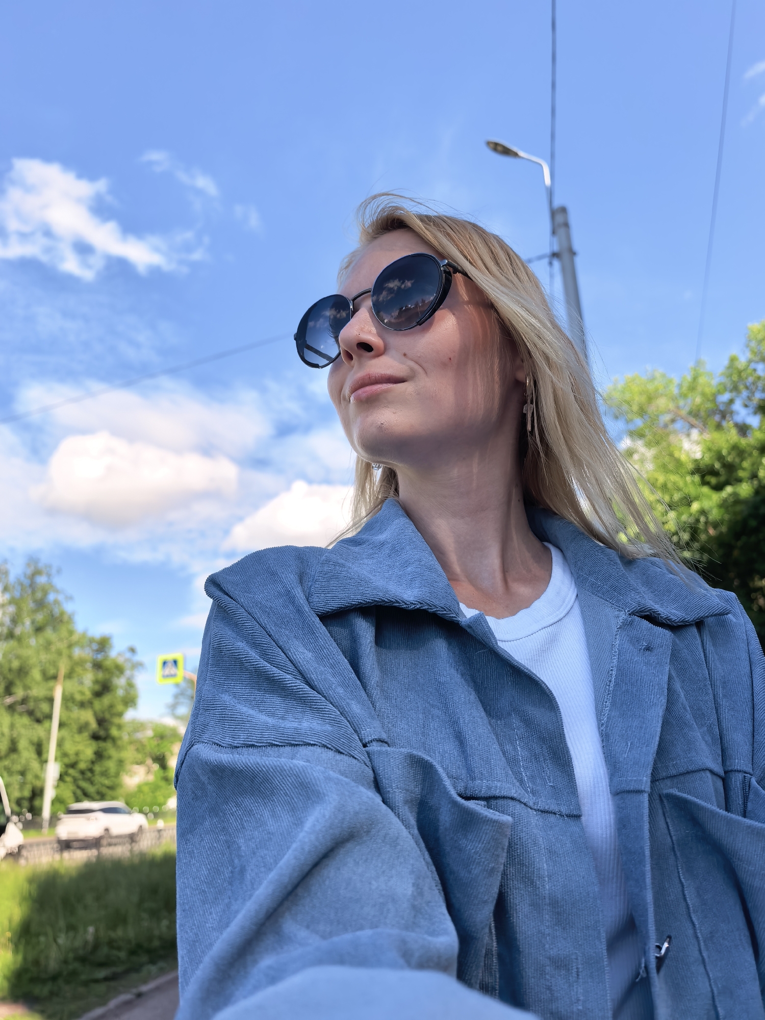 The image captures a blonde female from a low-angle perspective, looking slightly upward and to her right, wearing dark, round sunglasses that reflect white clouds and green foliage. She has long, straight blonde hair and is wearing a light blue, possibly corduroy or ribbed, oversized jacket over a white ribbed top, with gold-toned hoop earrings visible on her right ear.
The background reveals a bright blue sky with scattered white cumulus clouds. In the mid-ground, a paved road or sidewalk runs parallel to the viewer, flanked by green grass and lush trees. A yellow diamond-shaped road sign, indicating a pedestrian crossing, is visible on the left side, partially obscured by trees. A white SUV or similar vehicle is seen driving away in the distance on the road. On the right side, a tall, grey street light pole with a single light fixture extends upwards, accompanied by thin black utility lines stretching across the sky.
While the image is stated to be from the city of Sterlitamak, Russia, no specific landmarks, unique architecture, or distinctive street features are present within the visual evidence to forensically confirm a precise location within that city. The scene depicts a generic, tree-lined urban or suburban street environment under a clear sky.