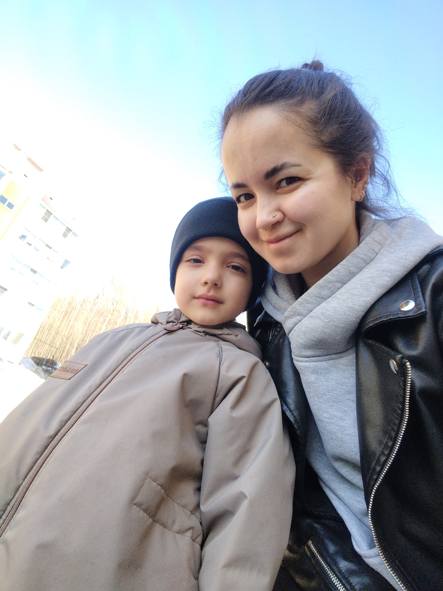 A selfie captures a woman and a child in an outdoor setting. The woman, on the right, wears a black leather jacket over a gray hoodie and has her dark hair tied back in a bun. She has a nose piercing and is smiling. The child, on the left, wears a dark beanie and a tan jacket with a hood. The background features a clear blue sky and a building with light-colored walls and multiple windows. Bare trees stand behind the child. The city of Noviy, Russia is mentioned as the location.