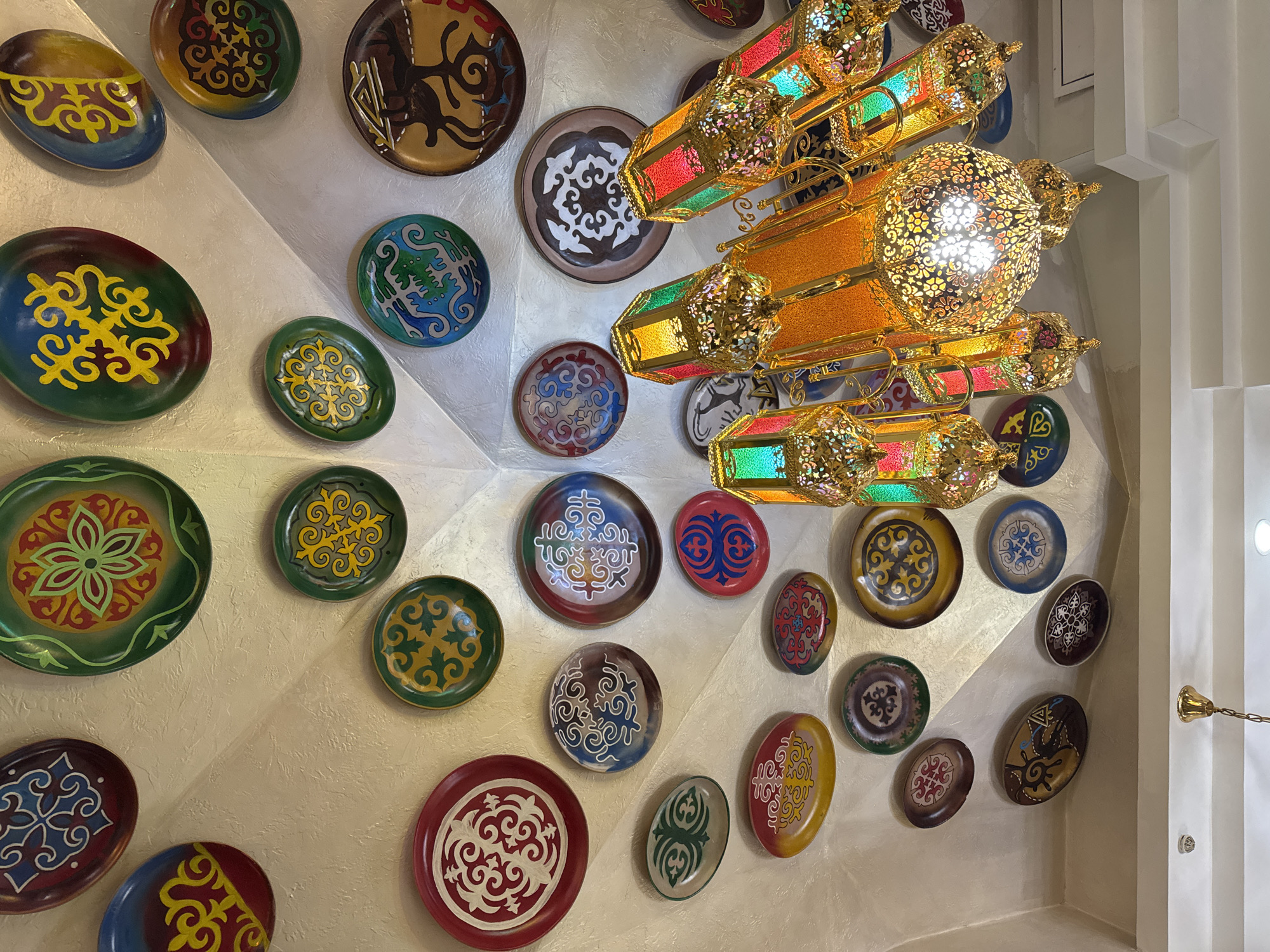 This image showcases a richly decorated interior wall adorned with numerous colorful, traditional plates and an elaborate chandelier. The setting suggests a public space like a restaurant, cafe, or hotel lobby, emphasizing a strong cultural aesthetic, consistent with Bishkek, Kyrgyzstan.
The wall features a textured, light-colored surface, possibly off-white or beige, and is constructed with various angles and segments, providing an dynamic backdrop for the display. Dozens of decorative plates of different sizes and shapes (round and oval) are meticulously arranged across these wall sections. These plates are hand-painted with intricate, stylized patterns characteristic of Central Asian or Kyrgyz ornaments. The motifs include swirling lines, geometric shapes, and stylized floral elements, presented in a vibrant palette of colors such as deep greens, blues, reds, yellows, browns, and whites.
A prominent, multi-tiered chandelier hangs from the ceiling in the upper right. It is crafted from golden, filigreed metal, forming multiple ornate lantern-like fixtures. Many of these individual lanterns incorporate colorful glass panels in shades of red, green, yellow, and blue, casting a warm, colored light. The central, spherical lights within the chandelier are brightly illuminated, creating intricate light patterns and highlighting the metallic details.
The overall impression is one of cultural richness and artistic craftsmanship. The lighting from the chandelier suggests an indoor setting, contributing to a warm and inviting ambiance. No specific activity is depicted, and there is no visible text.