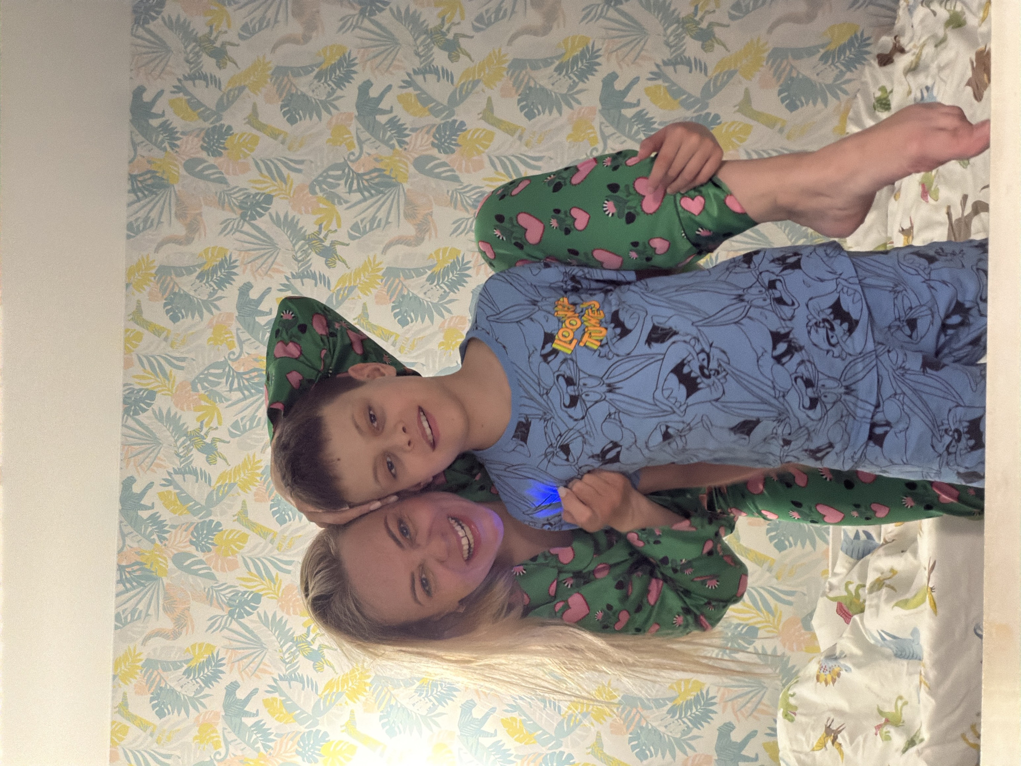 A medium shot captures a woman and a child, both smiling brightly and lying together, likely in a bedroom. The image is taken from an overhead perspective looking down at them.
The woman, with long blonde hair, is positioned lower in the frame, wearing green pajamas adorned with pink hearts and small green leaf motifs. She holds a small, blue glowing object in her hand. Her head is resting on the child's arm.
Above her, a child with short brown hair is also smiling. The child is wearing blue pajamas featuring cartoon characters, specifically Looney Tunes characters such as Bugs Bunny. A yellow patch on the child's chest clearly displays the text "LOONEY TUNES."
The background consists of patterned wallpaper behind them, featuring a light tropical design with leaves and stylized animals (possibly monkeys or chameleons) in shades of yellow, light blue, and pale pink/beige. To the right, a section of bedding with a different animal pattern (potentially dinosaurs) is visible.
Both individuals appear happy and relaxed, suggesting a casual and joyful moment, possibly in the morning or evening given their sleepwear. There are no specific cues within the image to confirm the location as Votkinsk, Russia.
