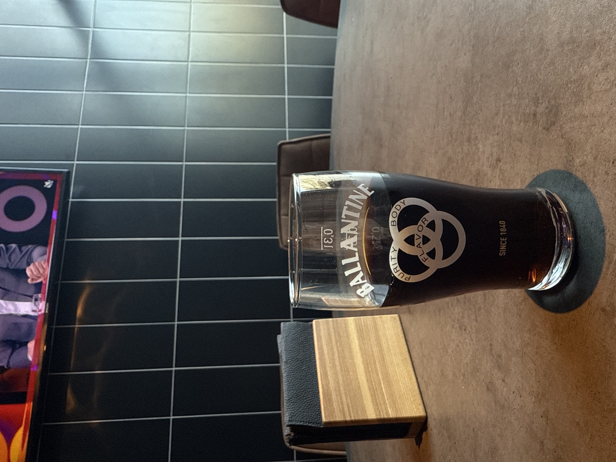The image shows a glass of dark liquid, likely a beer or stout, sitting on a black coaster atop a brown, textured table surface. Adjacent to the glass is a stack of small wooden and leather-bound books. The background consists of a wall covered in dark grey rectangular tiles. A portion of a television screen showing images of people is visible to the left of the wall. The image suggests an indoor location, possibly a bar or restaurant. There is no information in the image to determine the city of Perm, Russia.